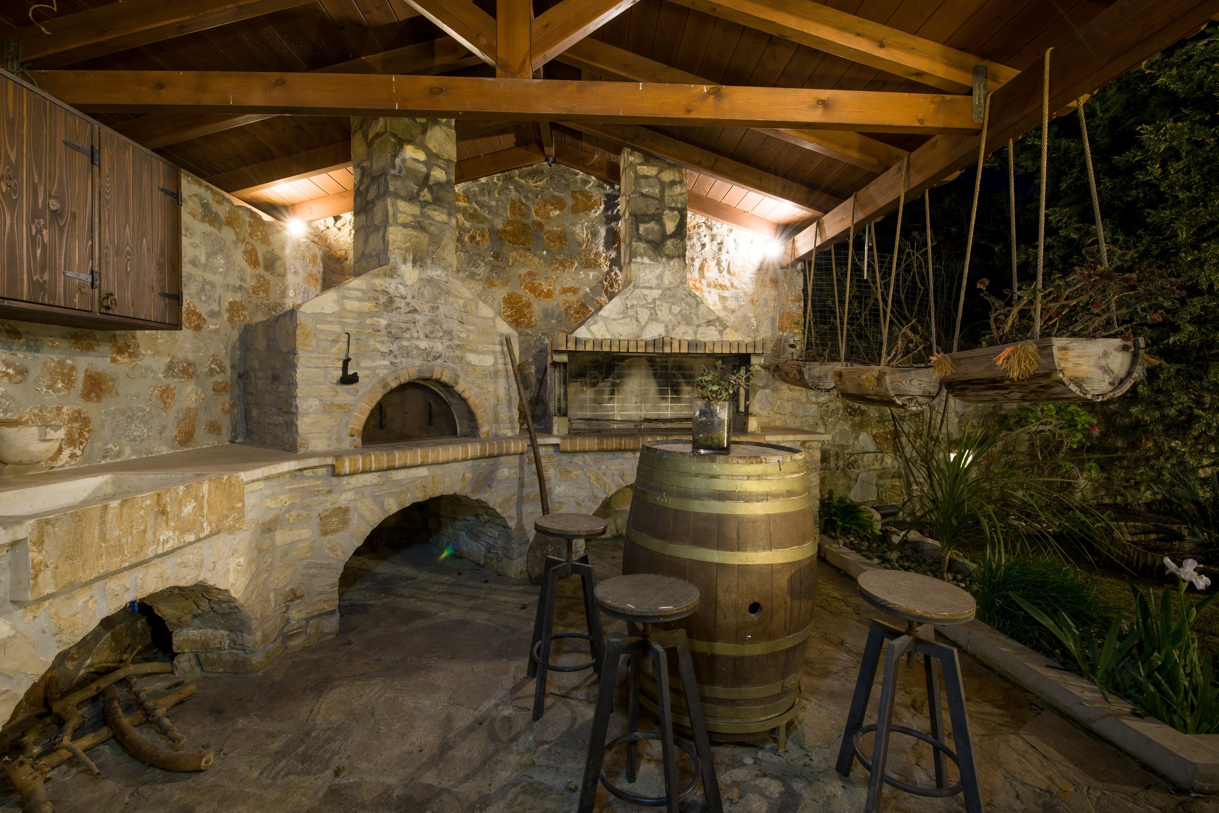 Outdoor rustic kitchen with stone oven, wooden barrel table, and stools under a wooden pergola.
