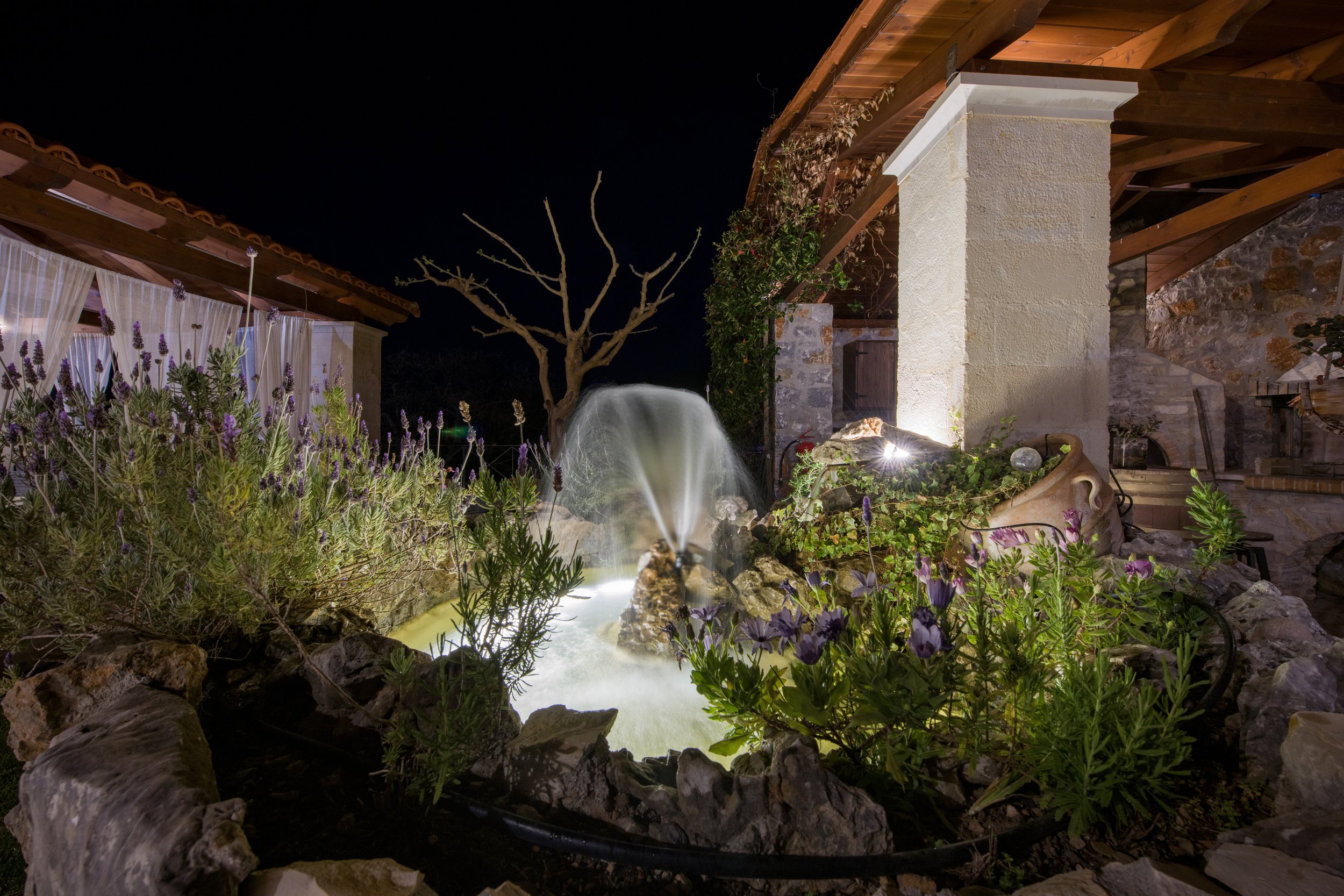 Nighttime garden with a small fountain, surrounded by stone and flowering plants, beside a wooden and stone building.