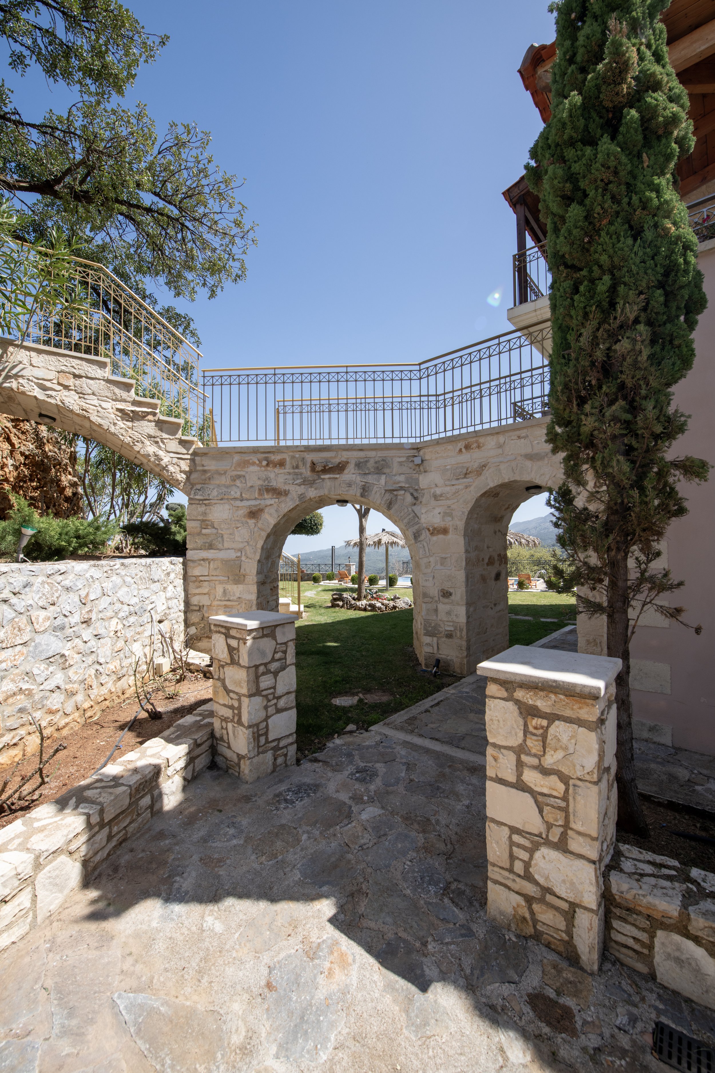 Stone archway and staircase with wrought iron railings, surrounded by a garden with trees and a clear blue sky.