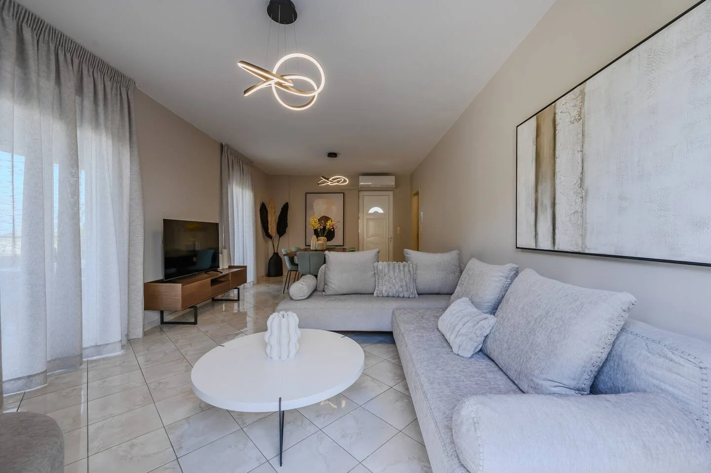 Living room with light-colored sofa, abstract wall art, white coffee table, TV on a wooden stand, and curtains on windows. Modern lighting fixtures hang from the ceiling.