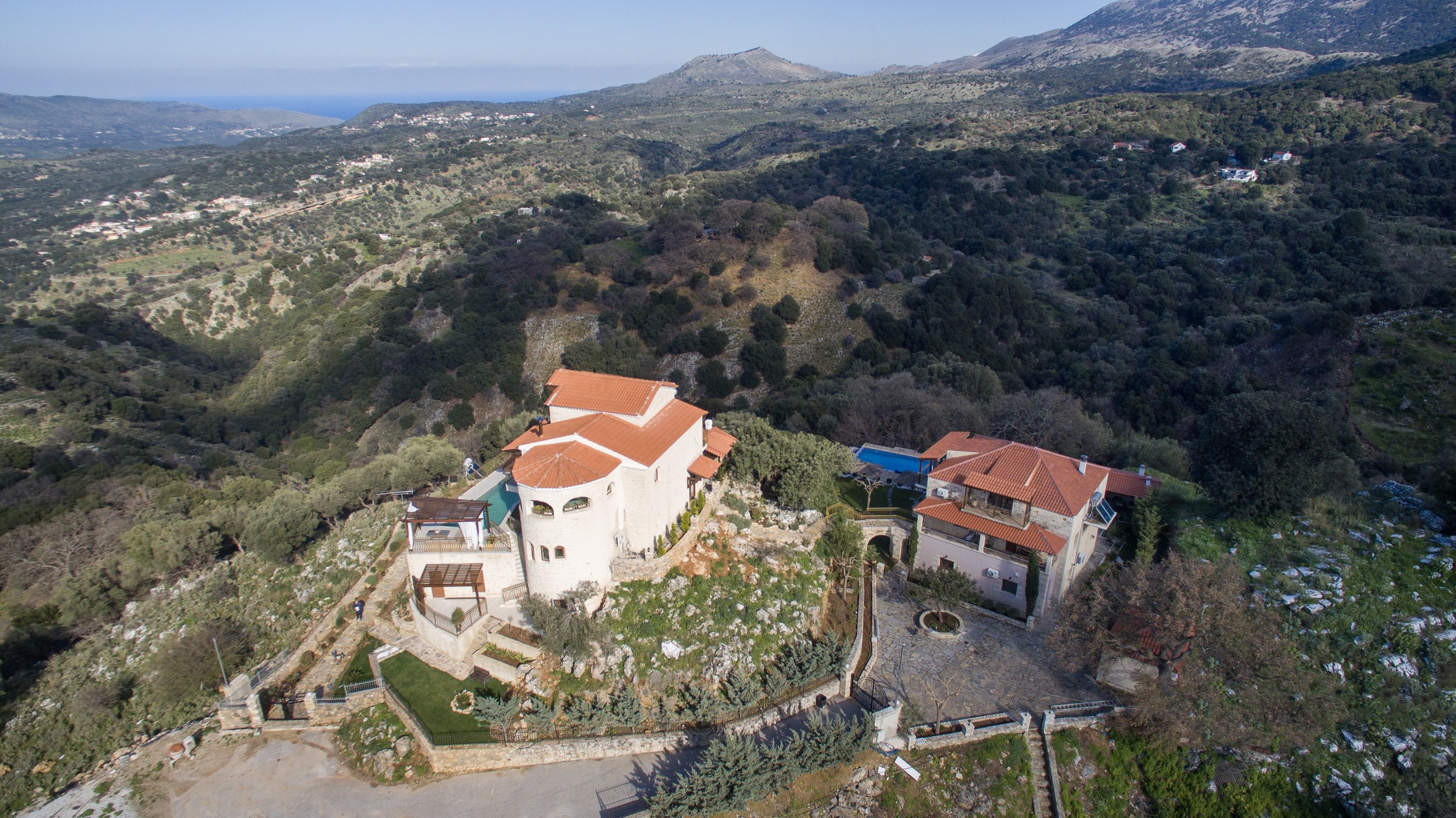 Aerial view of stone buildings with red roofs on a hilly landscape, surrounded by greenery and mountains. A pool is visible near one of the buildings. The area is rural and expansive, with few other structures in sight.