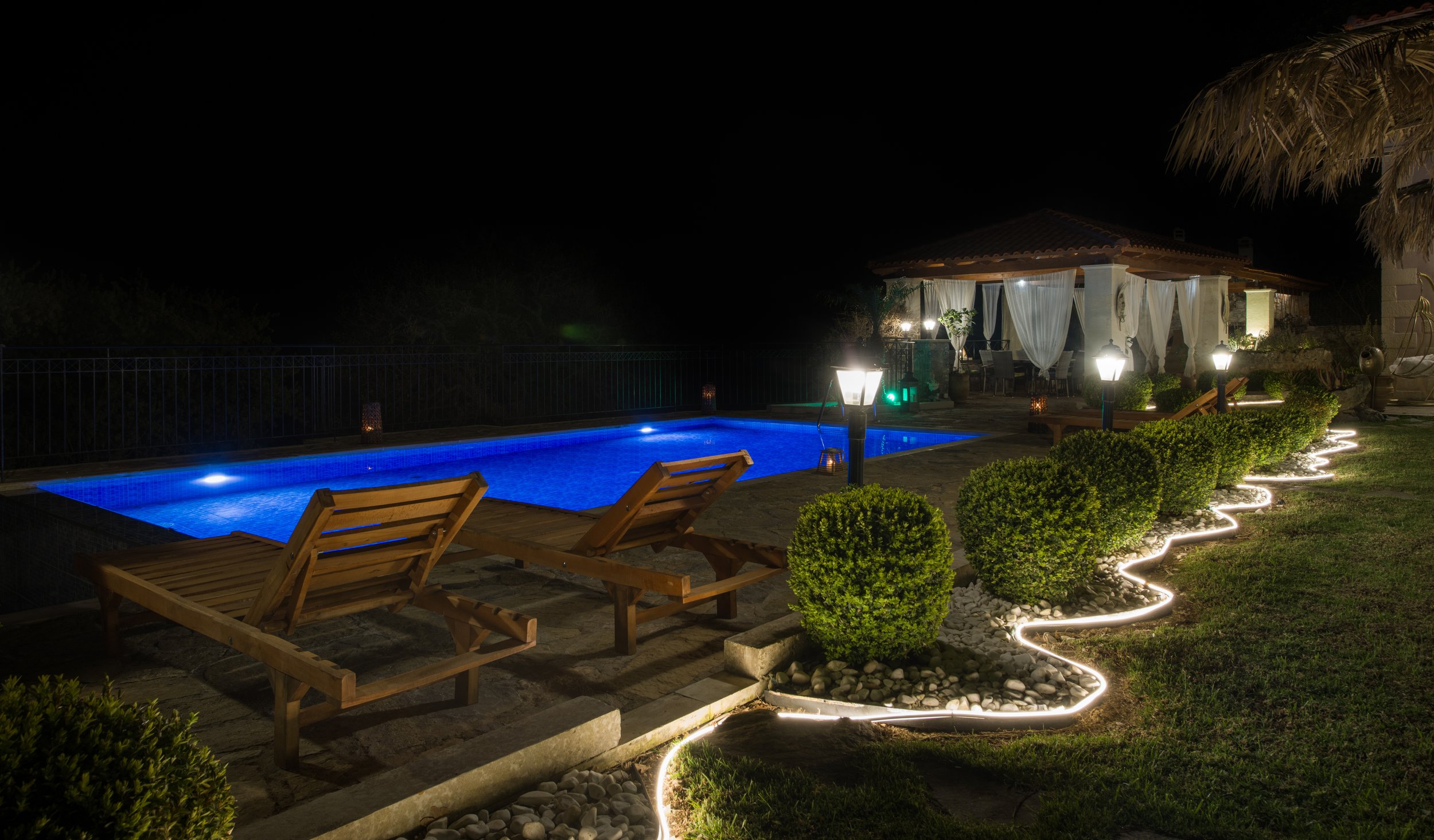 Nighttime view of a pool area with blue lights, surrounded by lounge chairs and landscaped greenery. A lit pathway and illuminated pavilion are visible in the background.