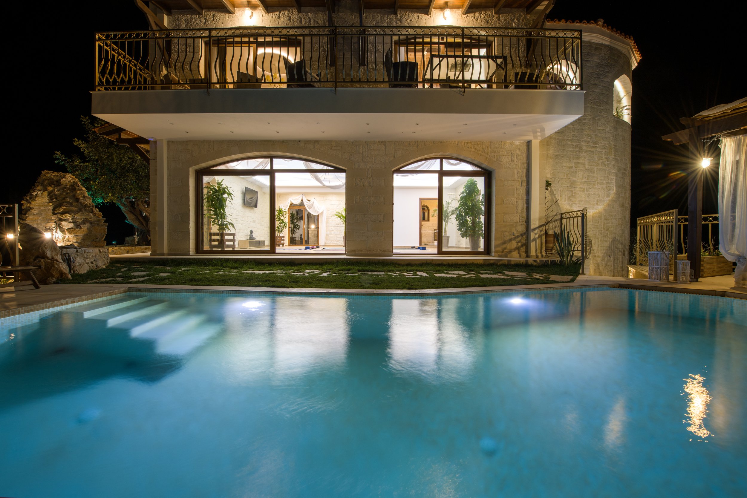 Luxury villa at night with illuminated swimming pool, large windows, and balcony.