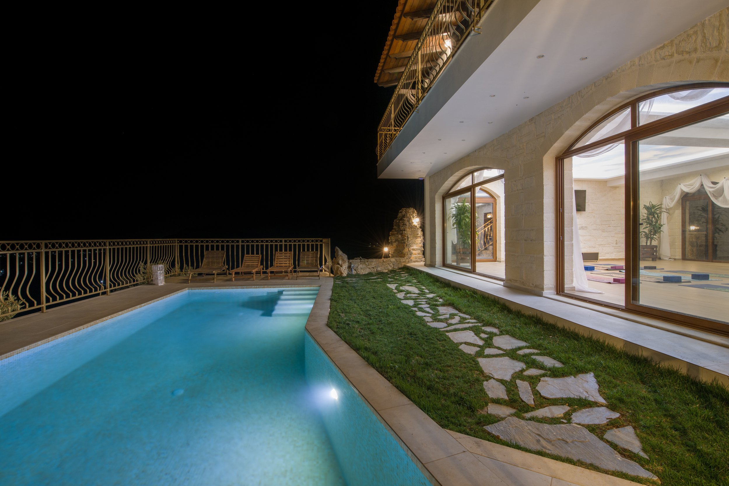 Night view of a luxury villa's swimming pool and patio with outdoor seating and large glass doors leading to the interior.