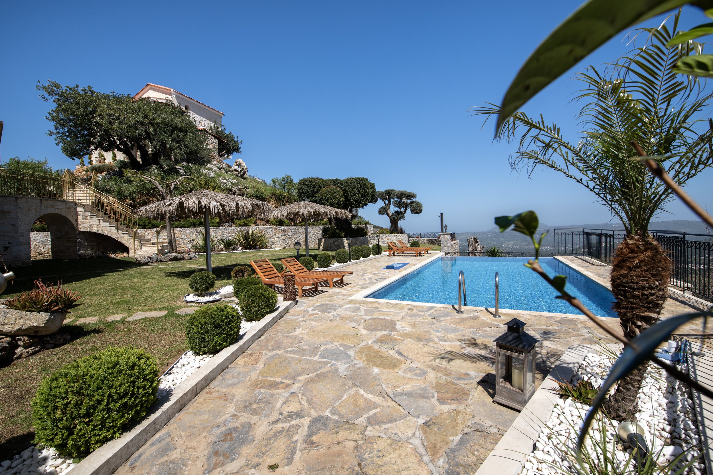 Outdoor scene with a swimming pool, wooden lounge chairs, and a stone patio surrounded by greenery and trees, under a clear blue sky.
