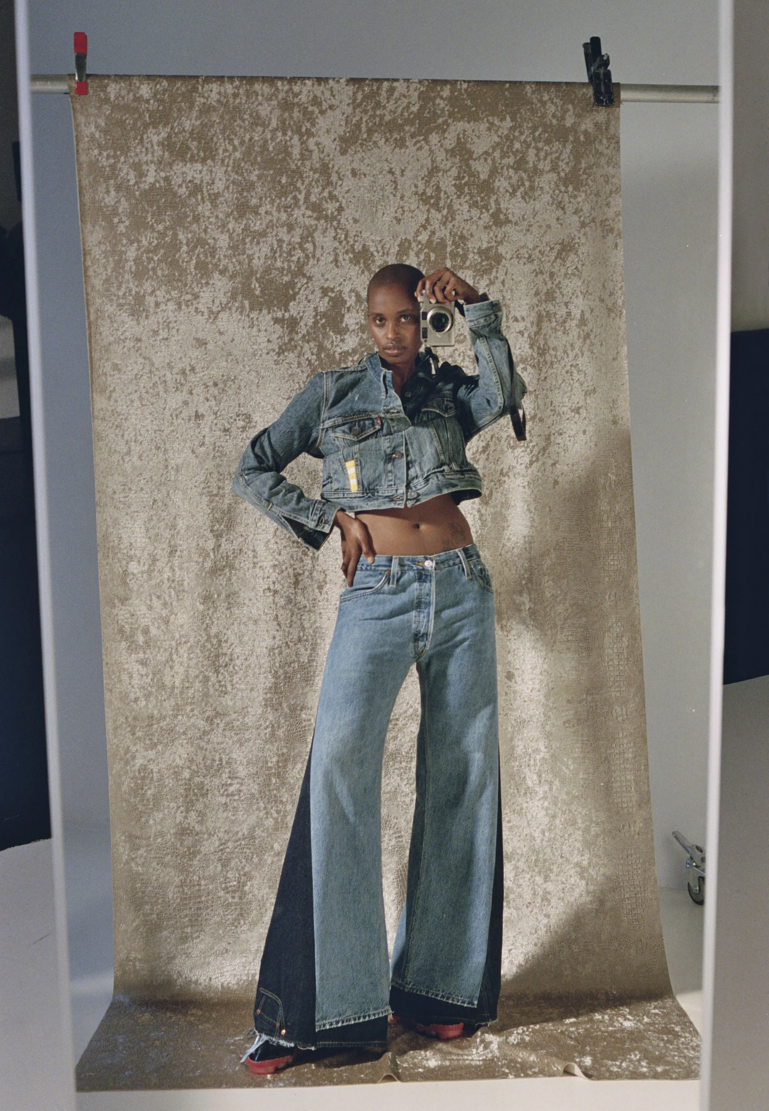 Person taking a mirror selfie in front of a textured beige backdrop, wearing a cropped denim jacket and high-waisted, flared jeans.