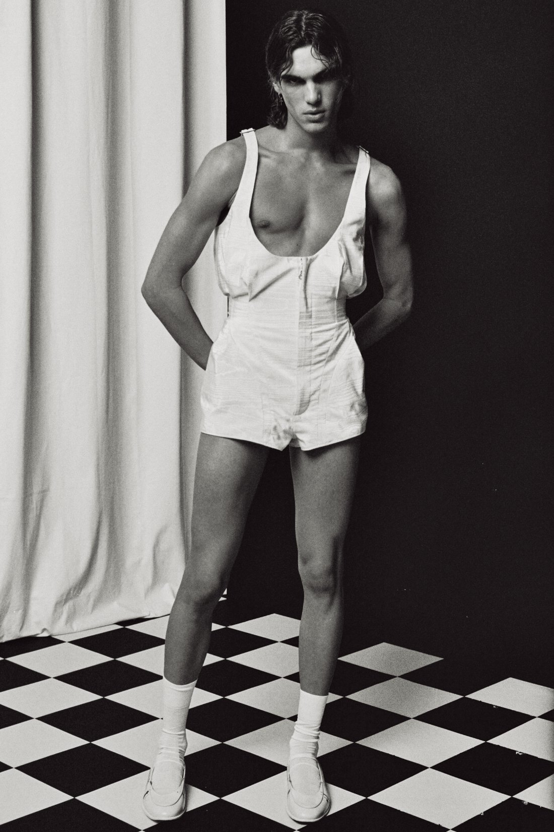 A person with short wavy hair wearing white shorts, socks, and shoes, standing with hands behind their back in front of a black and white checkered floor and black wall.