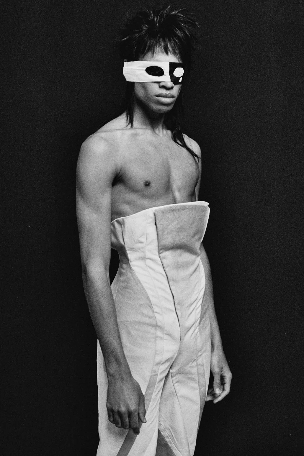 A person with short, spiked hair wearing a blindfold over their eyes, a strapless corset, and baggy pants against a dark background.