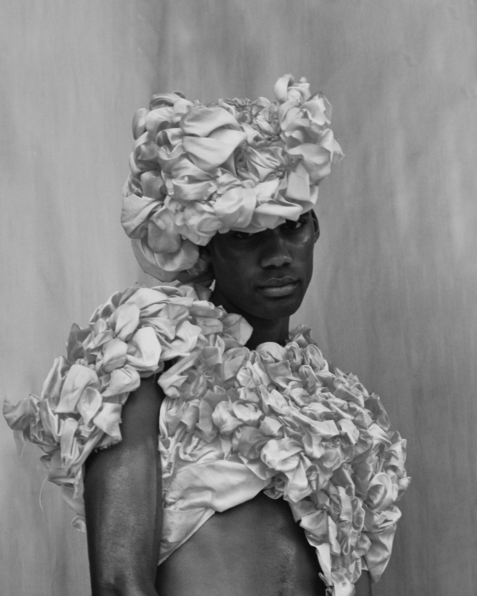 A person with dark skin wearing a large, textured headpiece, and matching shoulders and chest covering made of gathered fabric or ruffles, posed against a plain background.
