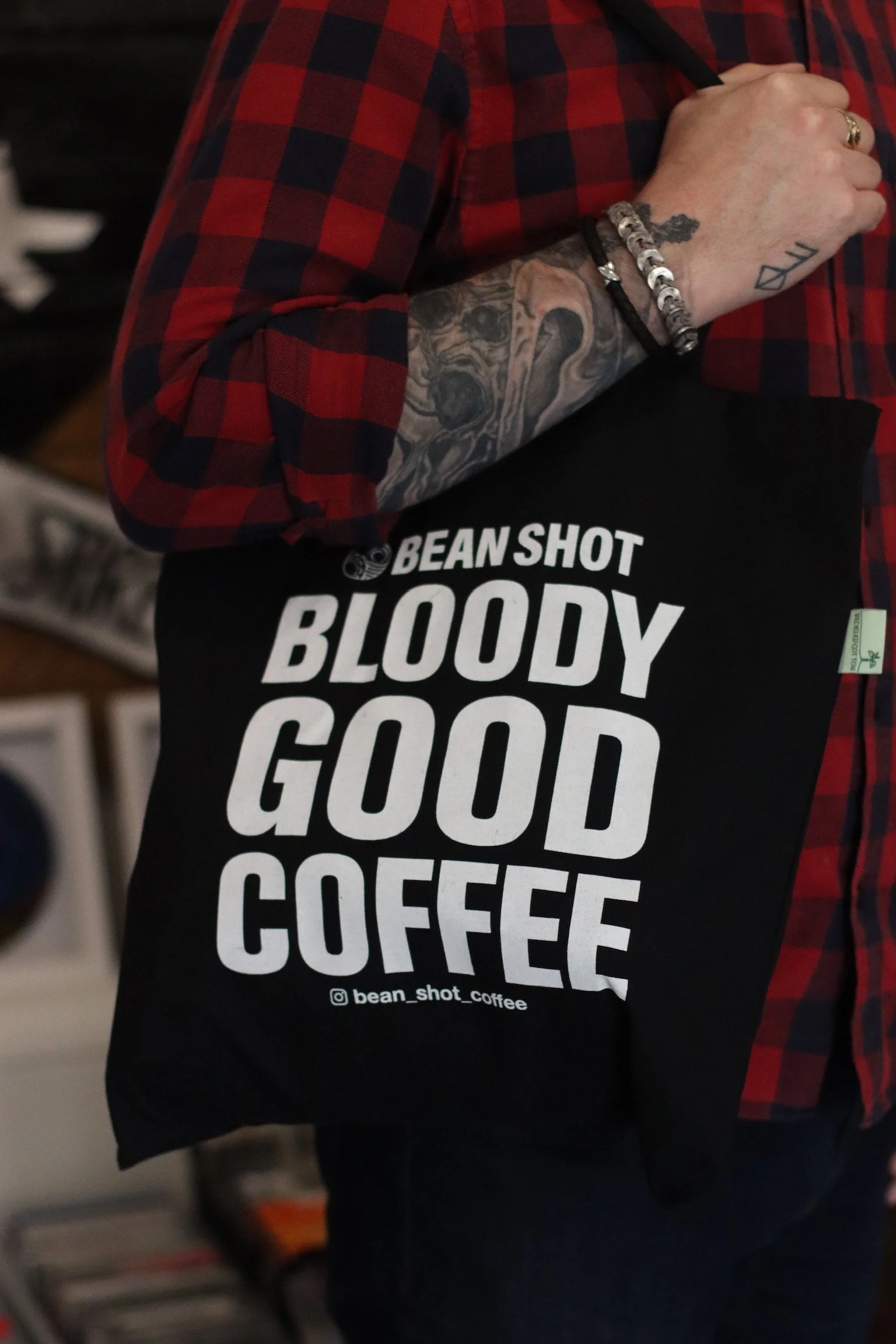 Bean shot bloody good coffee black tote bag being worn by someone wearing a red and black check shirt with arm tattoos shown