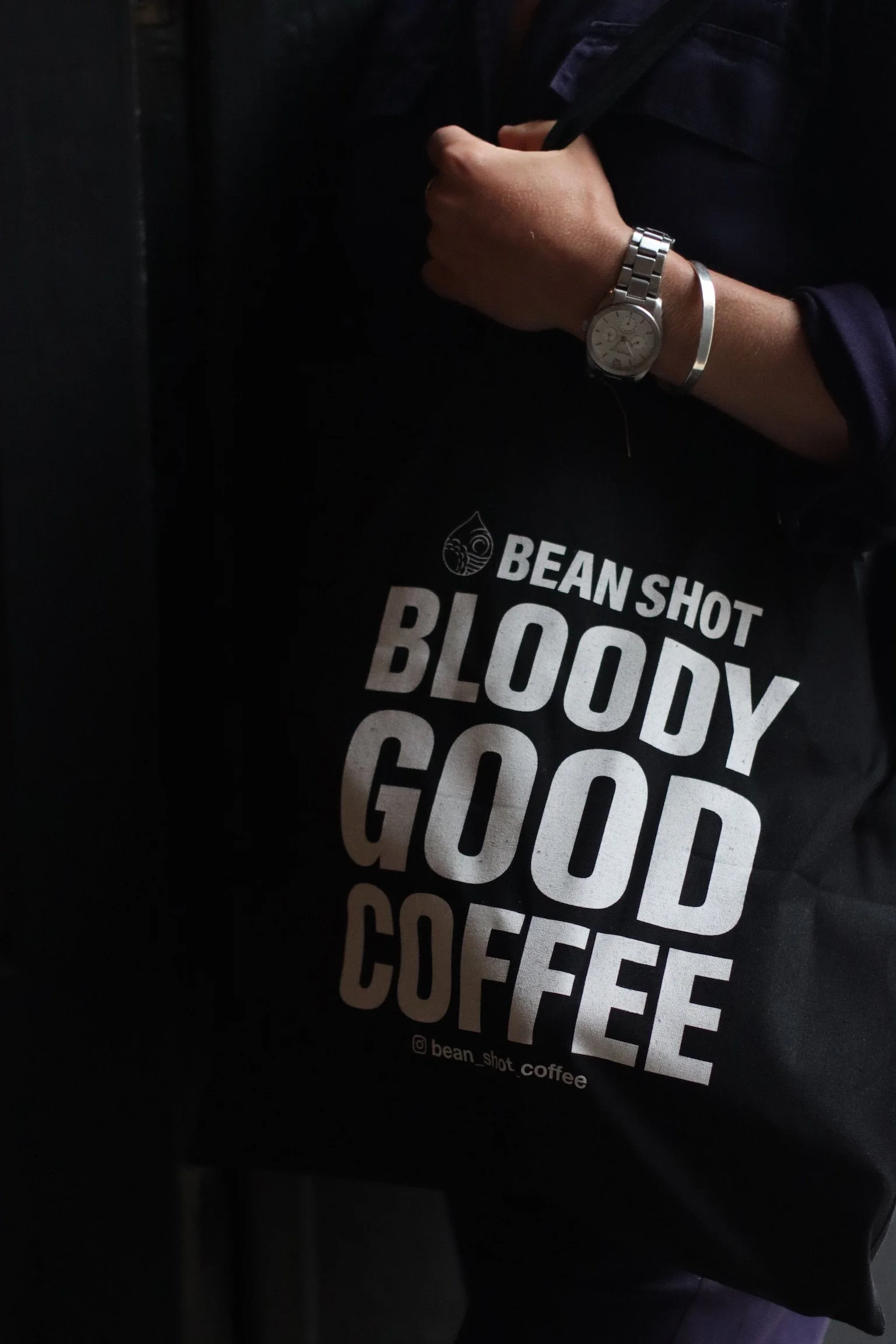 Bean shot bloody good coffee black tote bag being worn by someone wearing a silver watch and bracelet