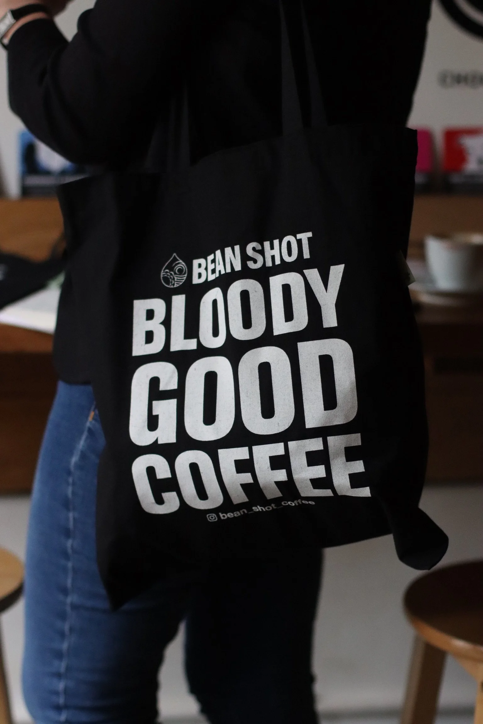 Bean shot bloody good coffee black tote bag being worn in coffee shop by someone wearing blue jeans