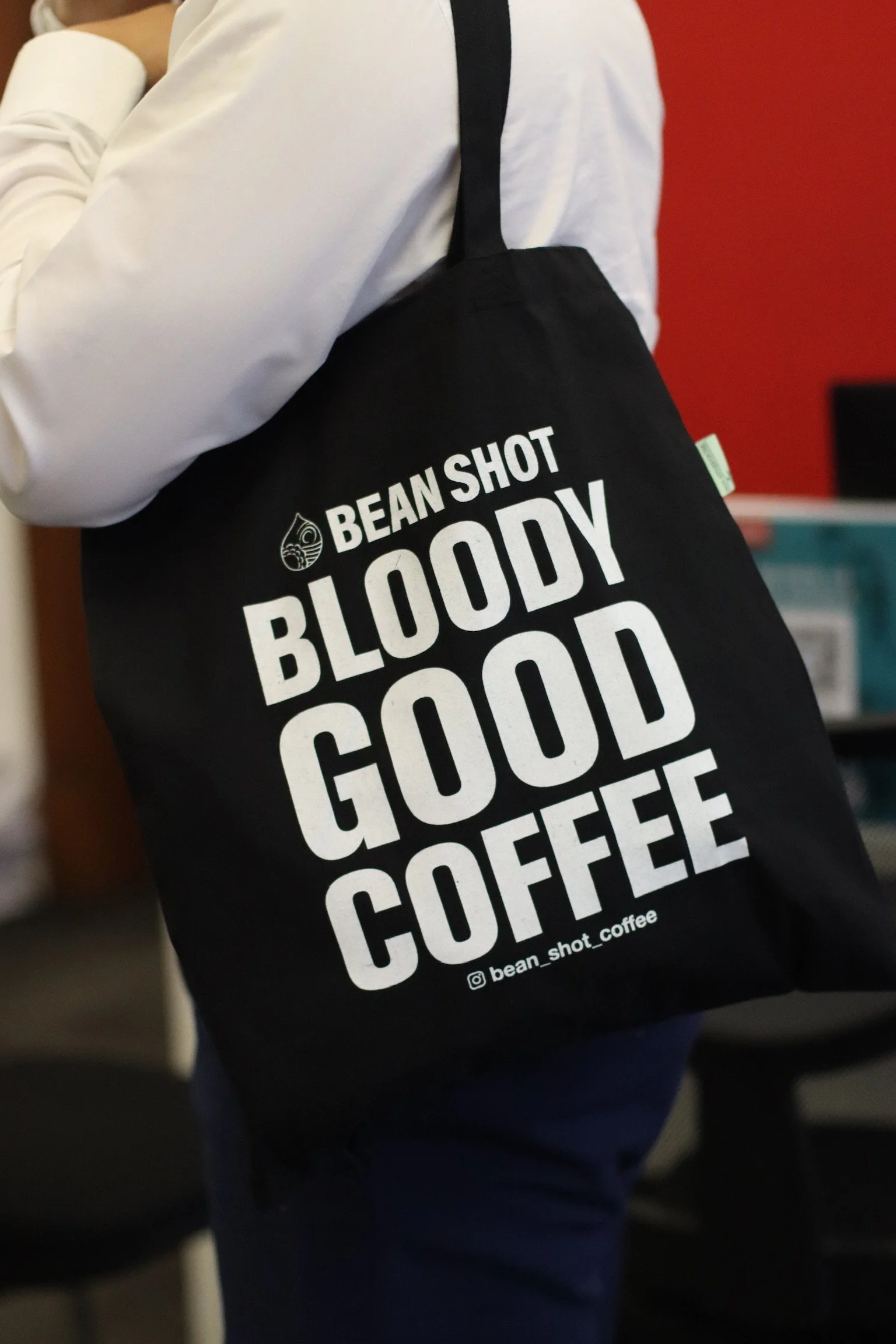 Bean shot bloody good coffee black tote bag being worn in coffee shop by someone wearing a white shirt with a red wall in the background