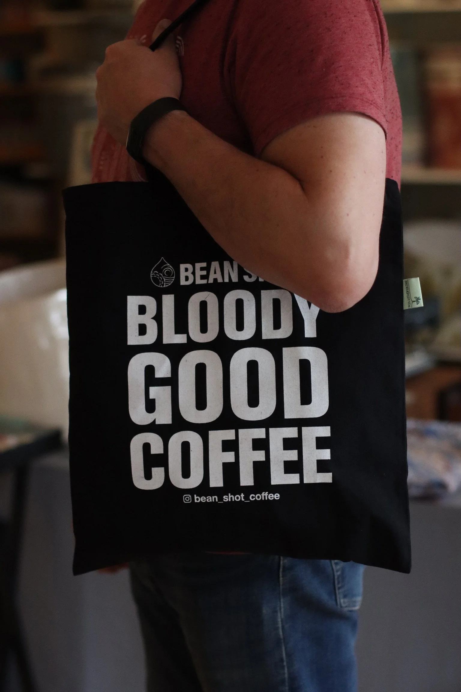 Bean shot bloody good coffee black tote bag being worn in coffee shop by someone wearing a red tshirt and black watch