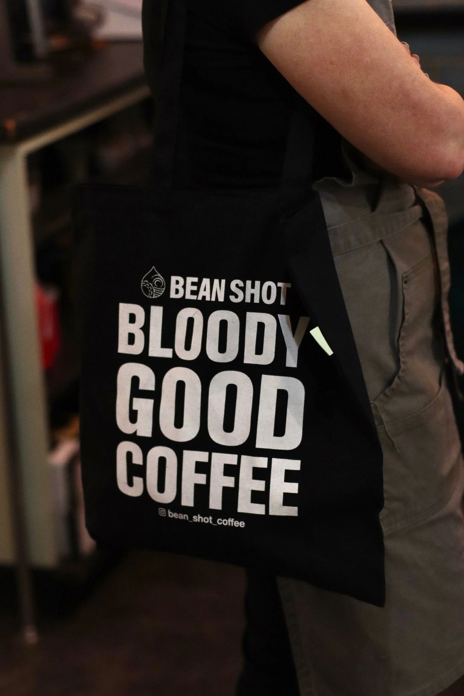 Bean shot bloody good coffee black tote bag being worn in coffee shop by someone wearing a grey apron