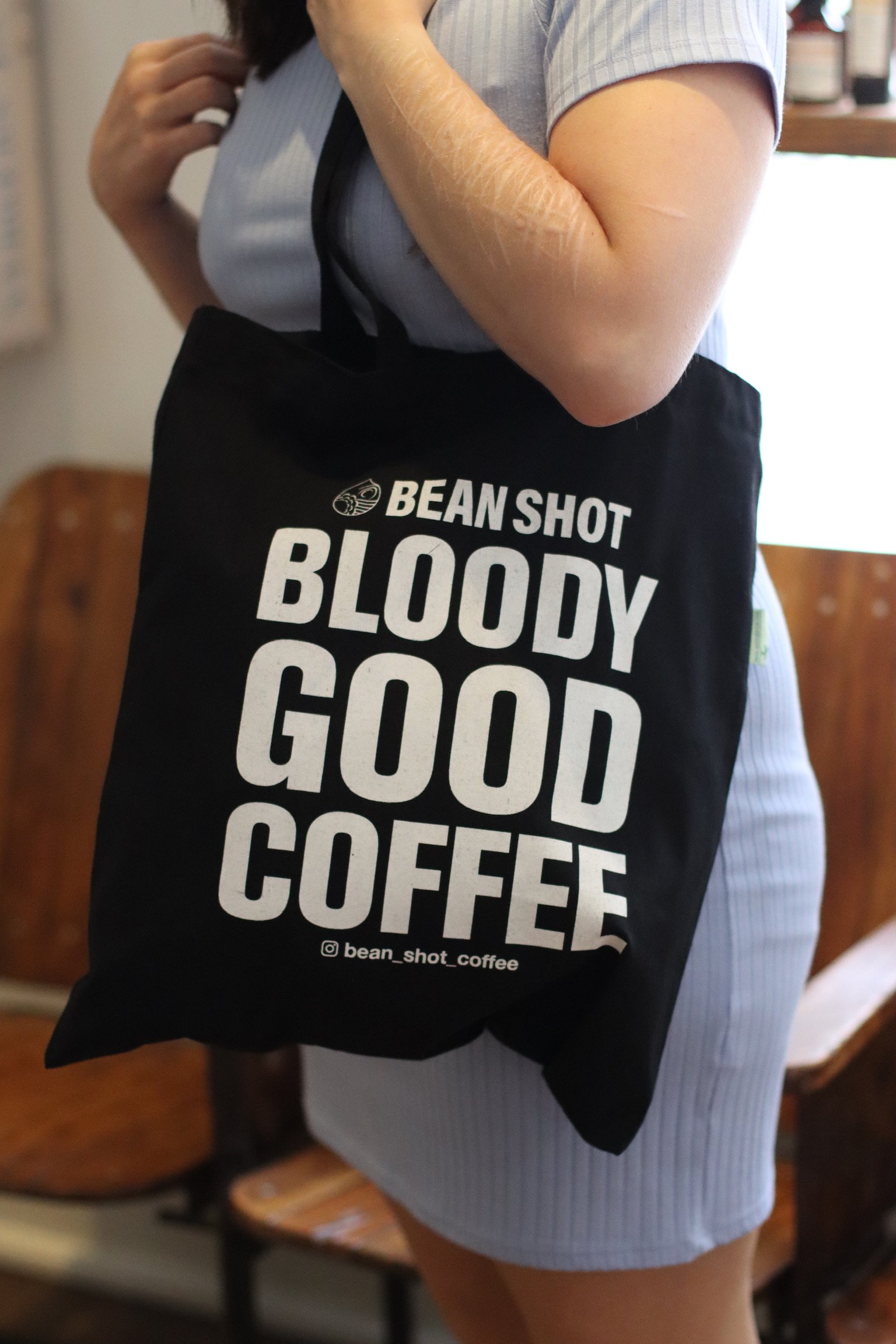 Bean shot bloody good coffee black tote bag being worn in coffee shop by someone wearing a light blue dress