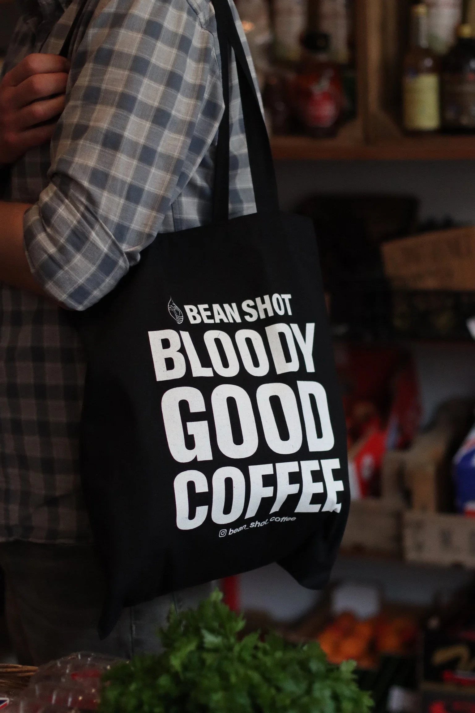 Bean shot bloody good coffee black tote bag being worn in fruit and vegetable shop by someone wearing a light blue grey check shirt