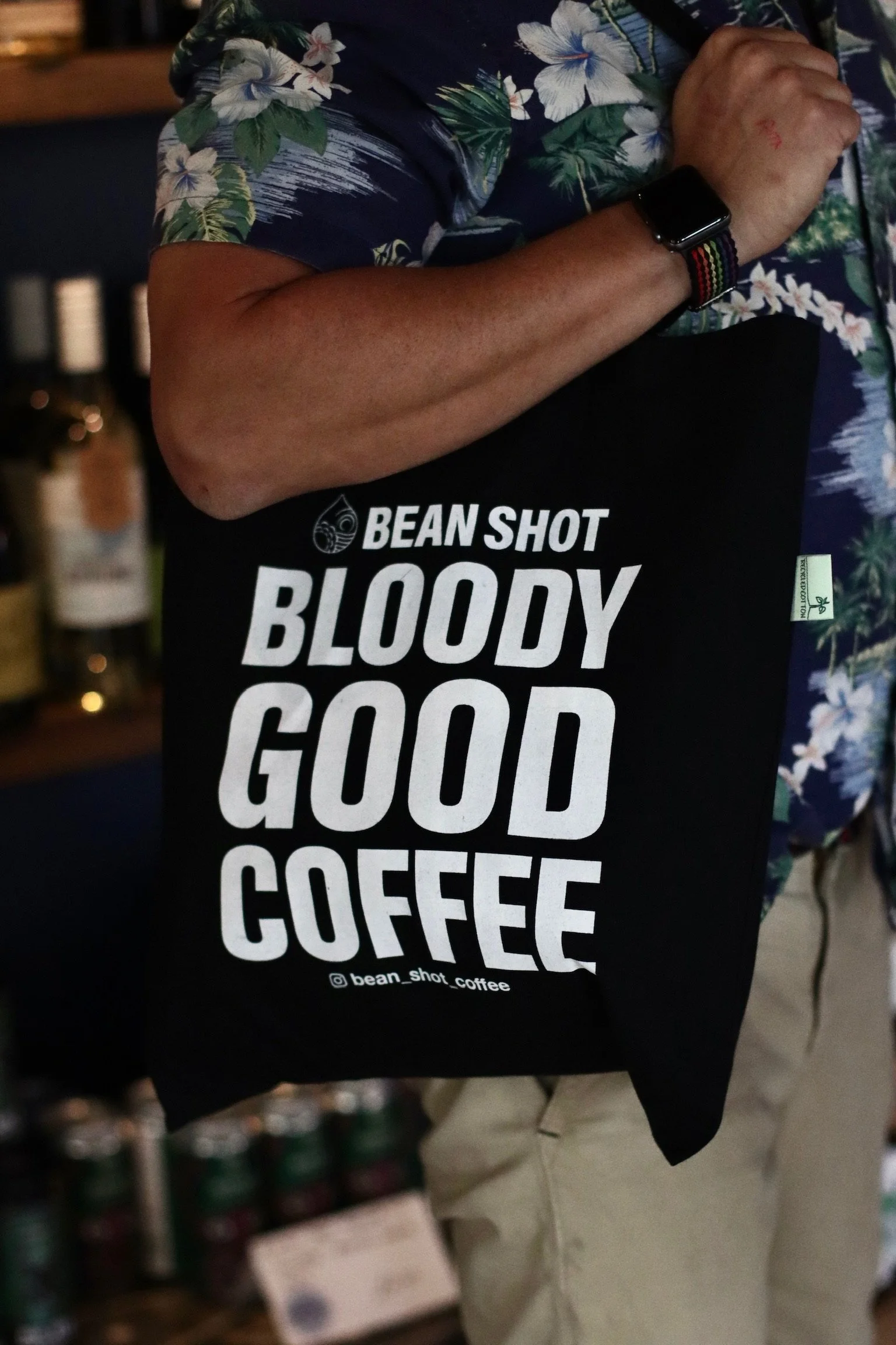 Bean shot bloody good coffee black tote bag being worn in wine shop by someone wearing a floral shirt and light shorts