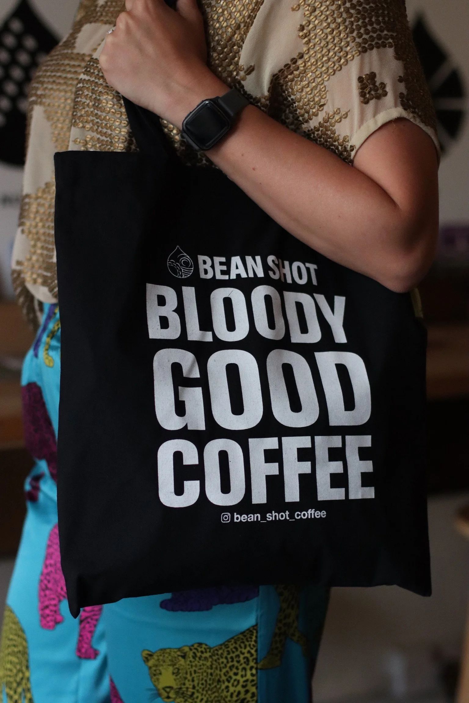 Bean shot bloody good coffee black tote bag being worn in coffee shop by someone wearing a blue animal print trousers and gold sequin top