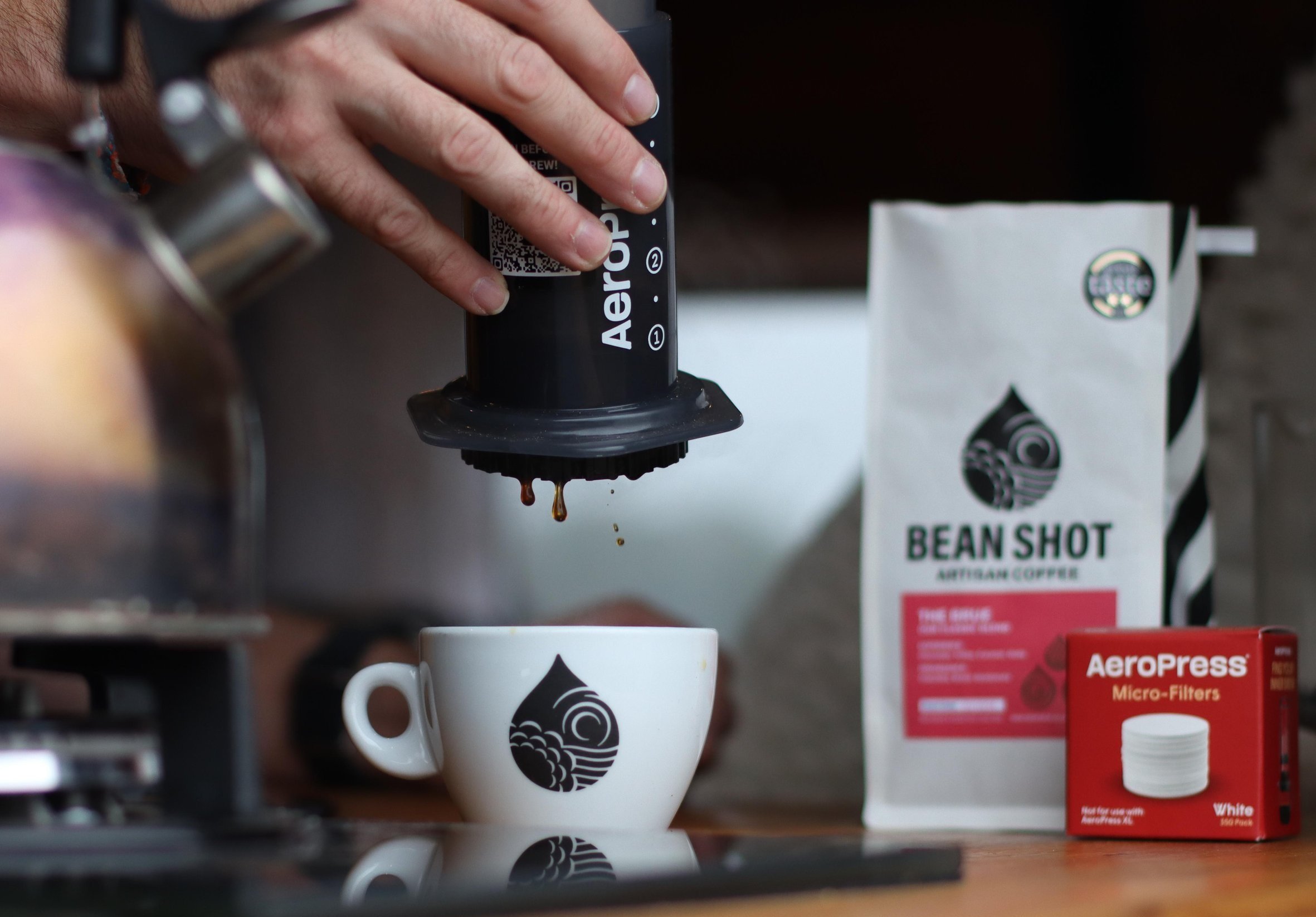 aeropress shown in use with coffee dripping into white ben shot cup. bag of bean shot coffee and box of aeropress filters shown in background .