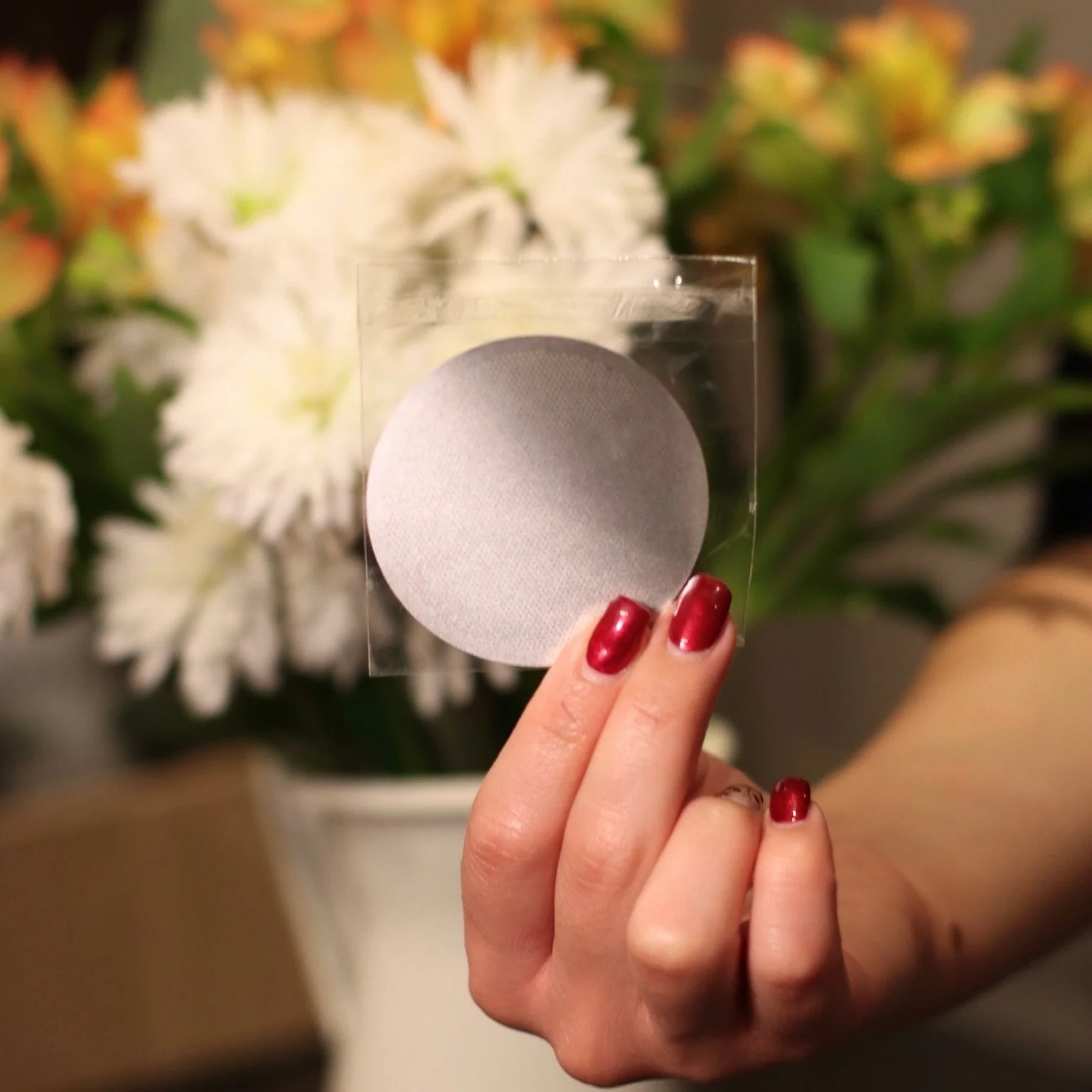Hand with red nails holding reusable metal aeropress filter in front of a vase of white and yellow flowers