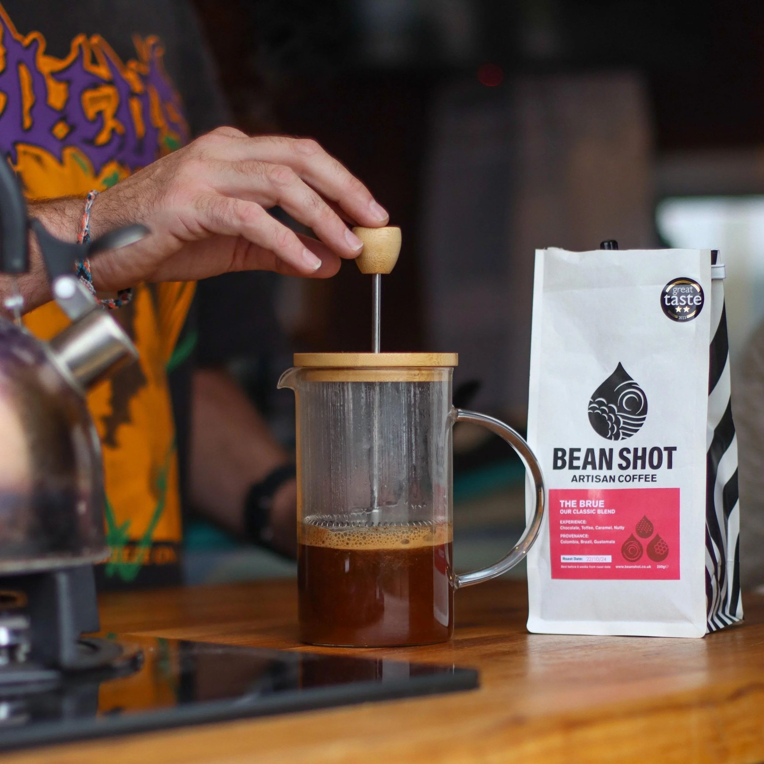 The Brue Bean Shot Coffee Beans in bag on wooden counter with a hand pressing down on a cafetiere making coffee