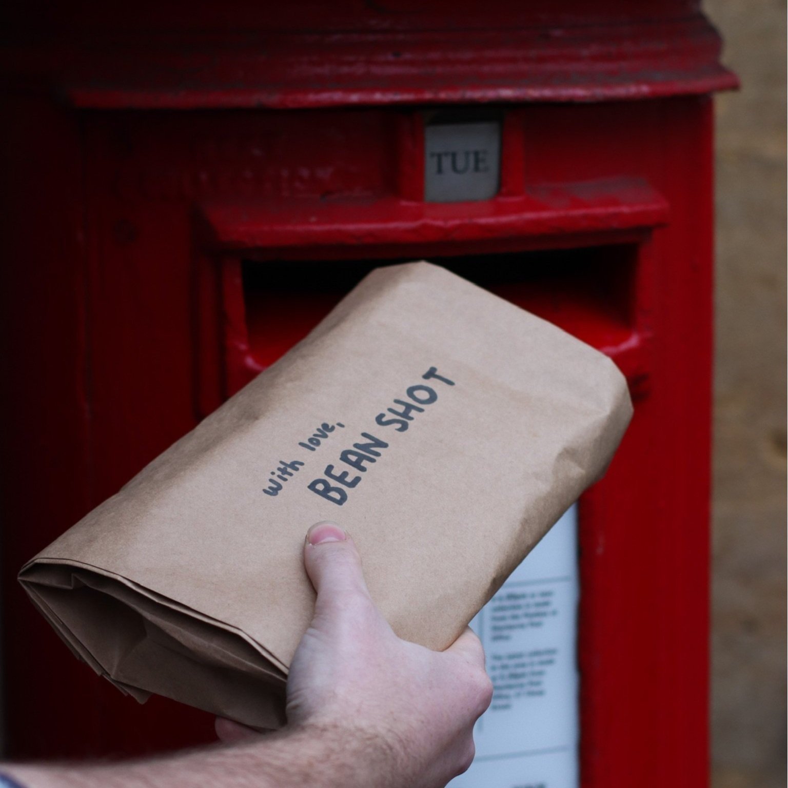 Brown paper parcel with black writing on that says with love from bean shot being posted by hand into a red post box.