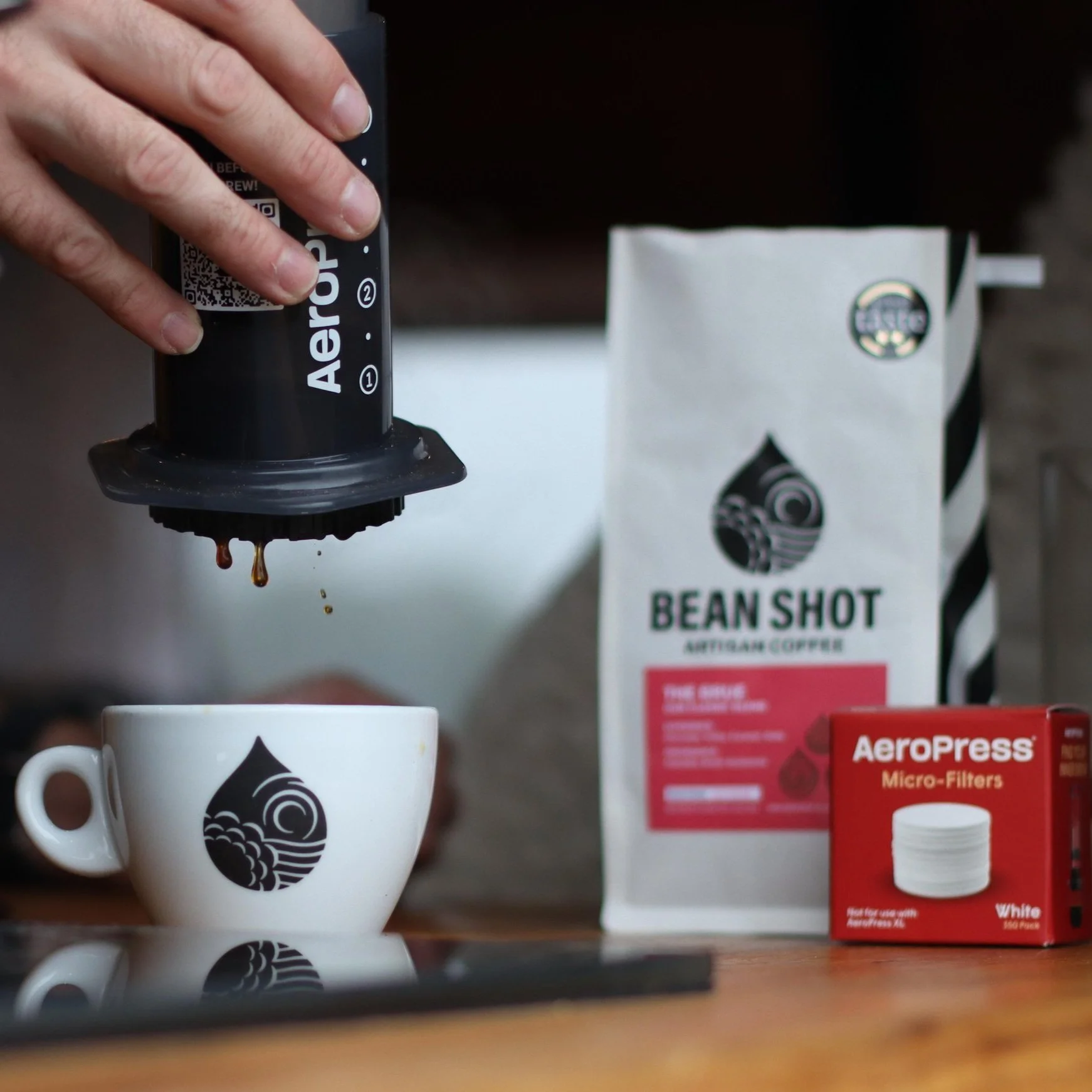 Aeropress being used and showing coffee dripping from device into white bean shot cup. Cup and aeropress shown on hob on wooden counter with bag of bean shot coffee and filters to the right