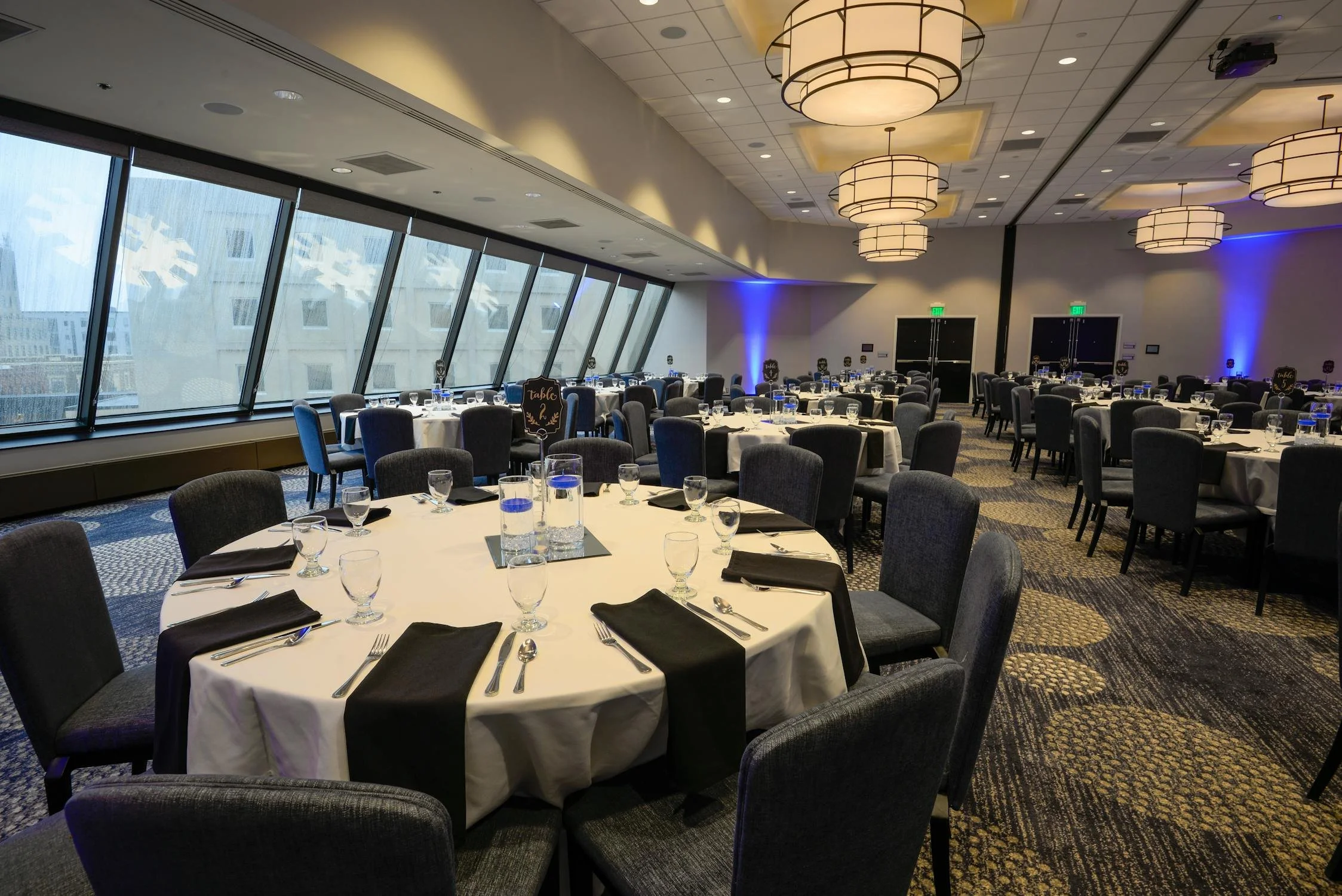 Elegant banquet hall with round tables, set with white tablecloths and black napkins, dark chairs, modern lighting, and large angled windows.