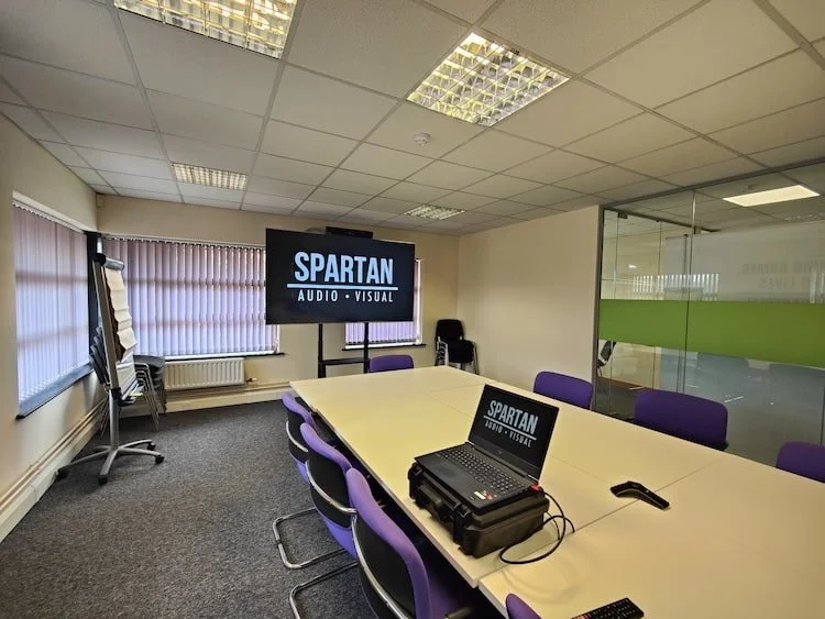 Modern conference room with a large monitor displaying 'Spartan Audio Visual,' a laptop on the table, purple chairs, and a flip chart.