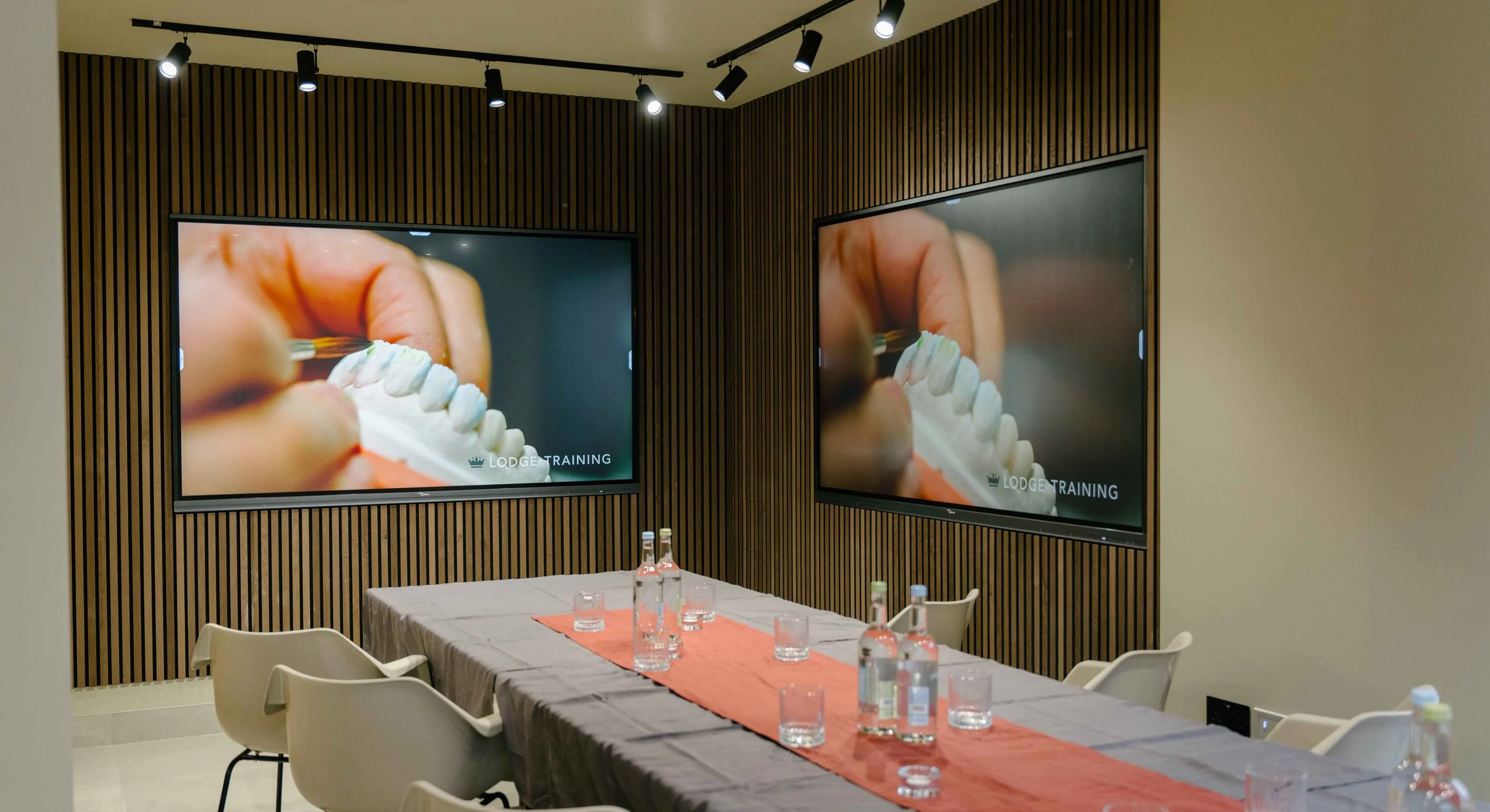 Conference room with two large screens displaying close-up images of hands painting dental molds, with "Lodge Training" text. A table with bottled water and glasses is set in the room.