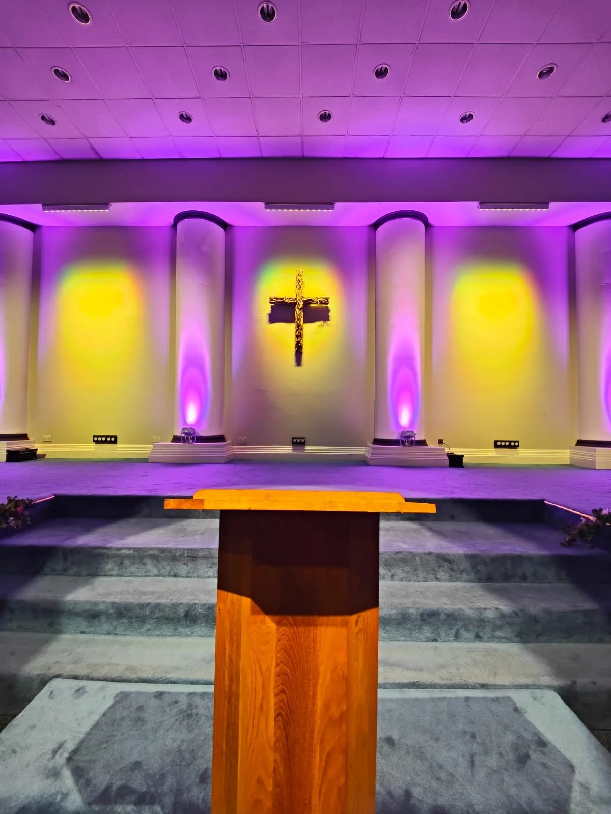 Church interior with wooden podium, cross on wall, purple and yellow lighting