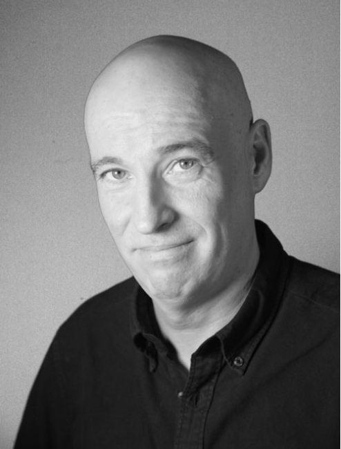 Black and white portrait of a bald man wearing a dark shirt, looking at the camera with a slight smile.