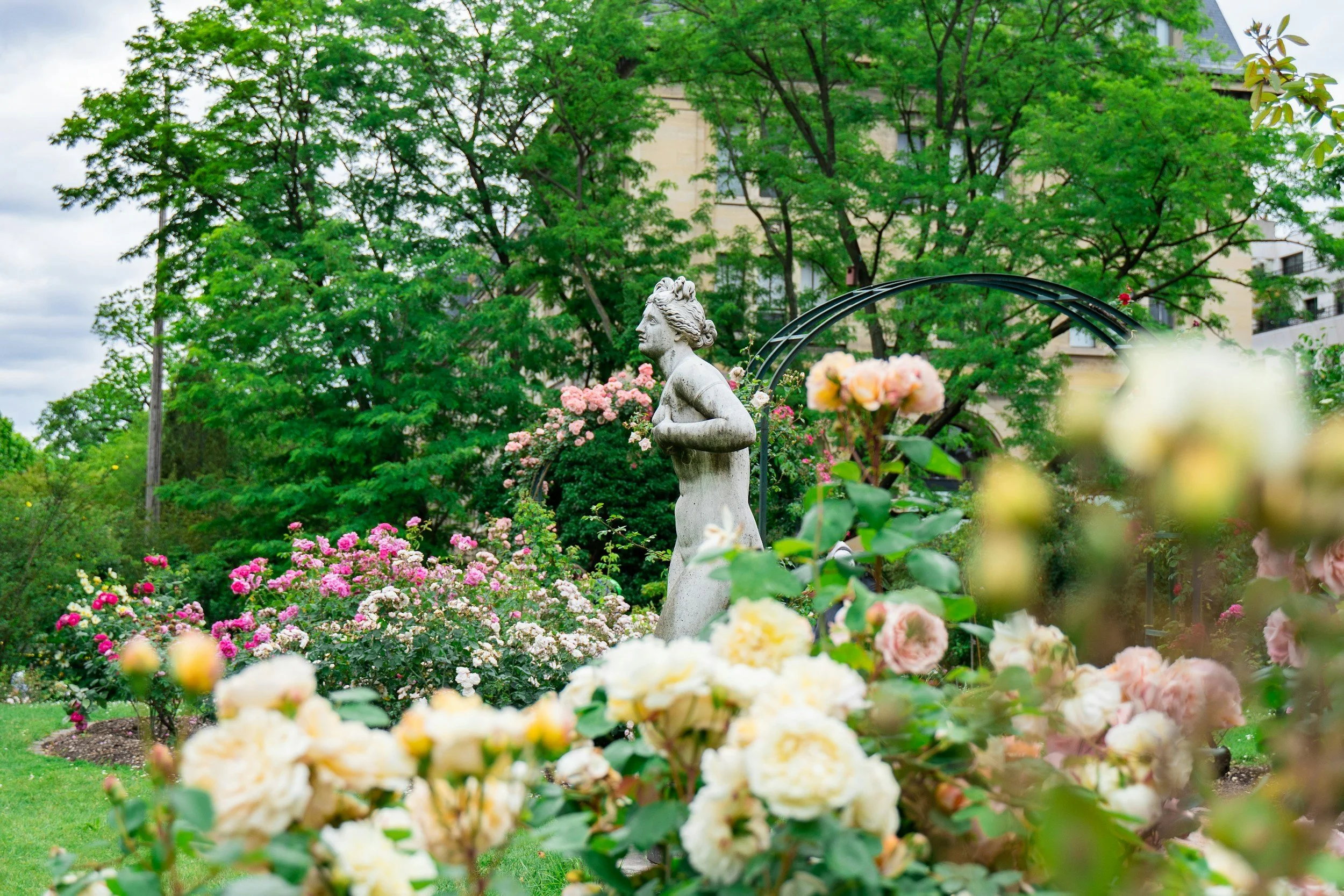 A garden with a statue of a woman surrounded by colorful pink, white, and yellow roses and green trees in the background.