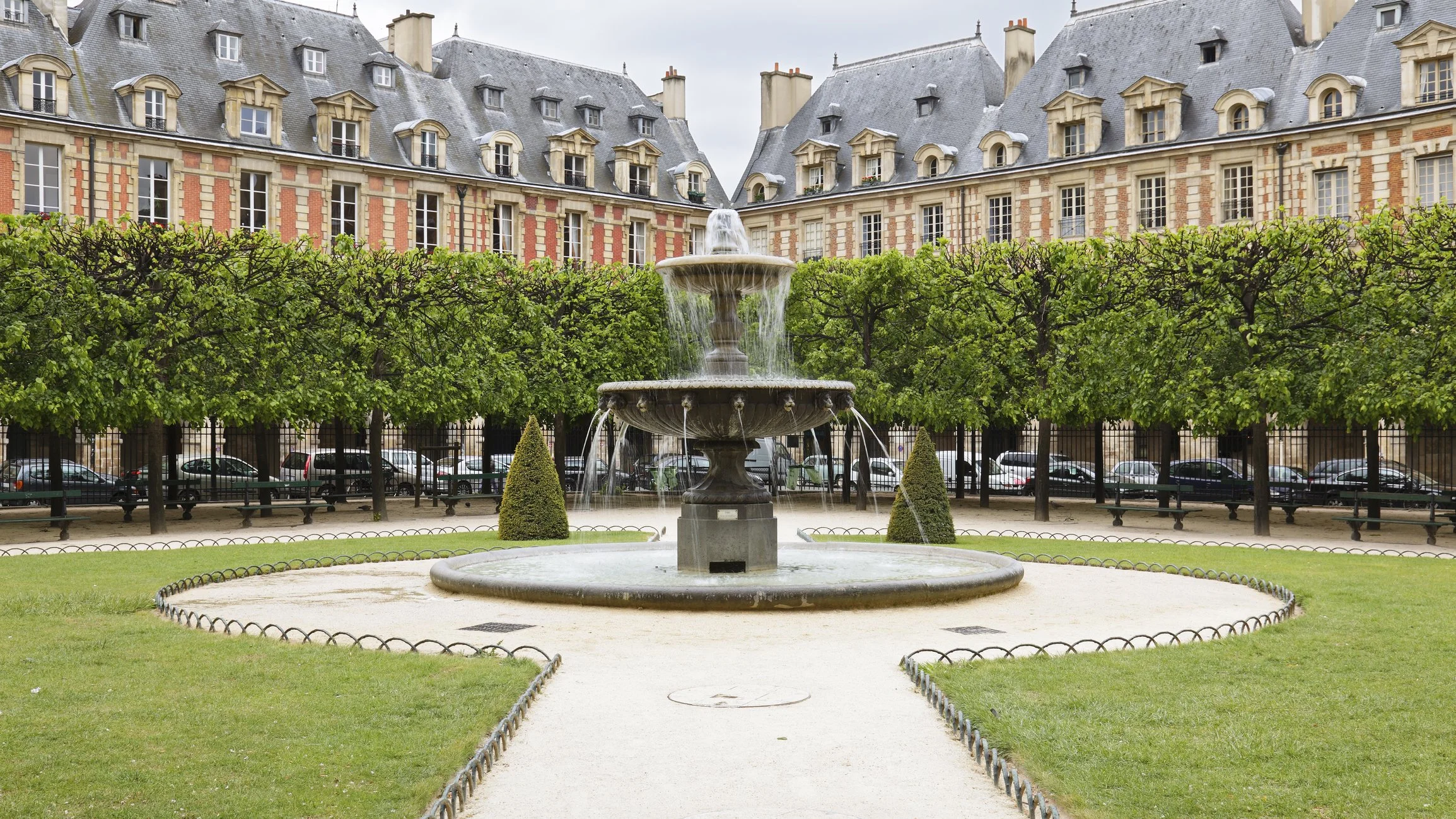 A city park with a fountain and trimmed bushes, surrounded by a wrought iron fence and classic European-style buildings with gray mansard roofs in the background.