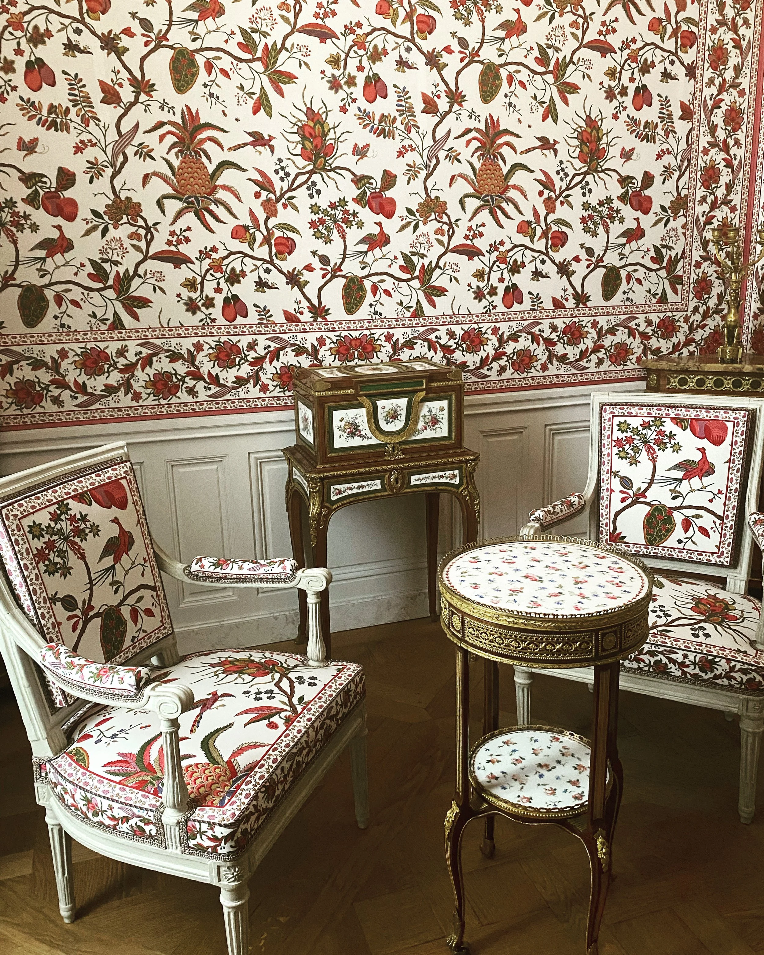 A historic library with floor-to-ceiling wooden bookshelves, some with glass doors, filled with red and brown books. A marble-topped window sill runs along the right wall, and a large crystal chandelier hangs from the ceiling, illuminating the room. The room has a parquet wooden floor with a geometric pattern and green drapery behind the bookshelves.