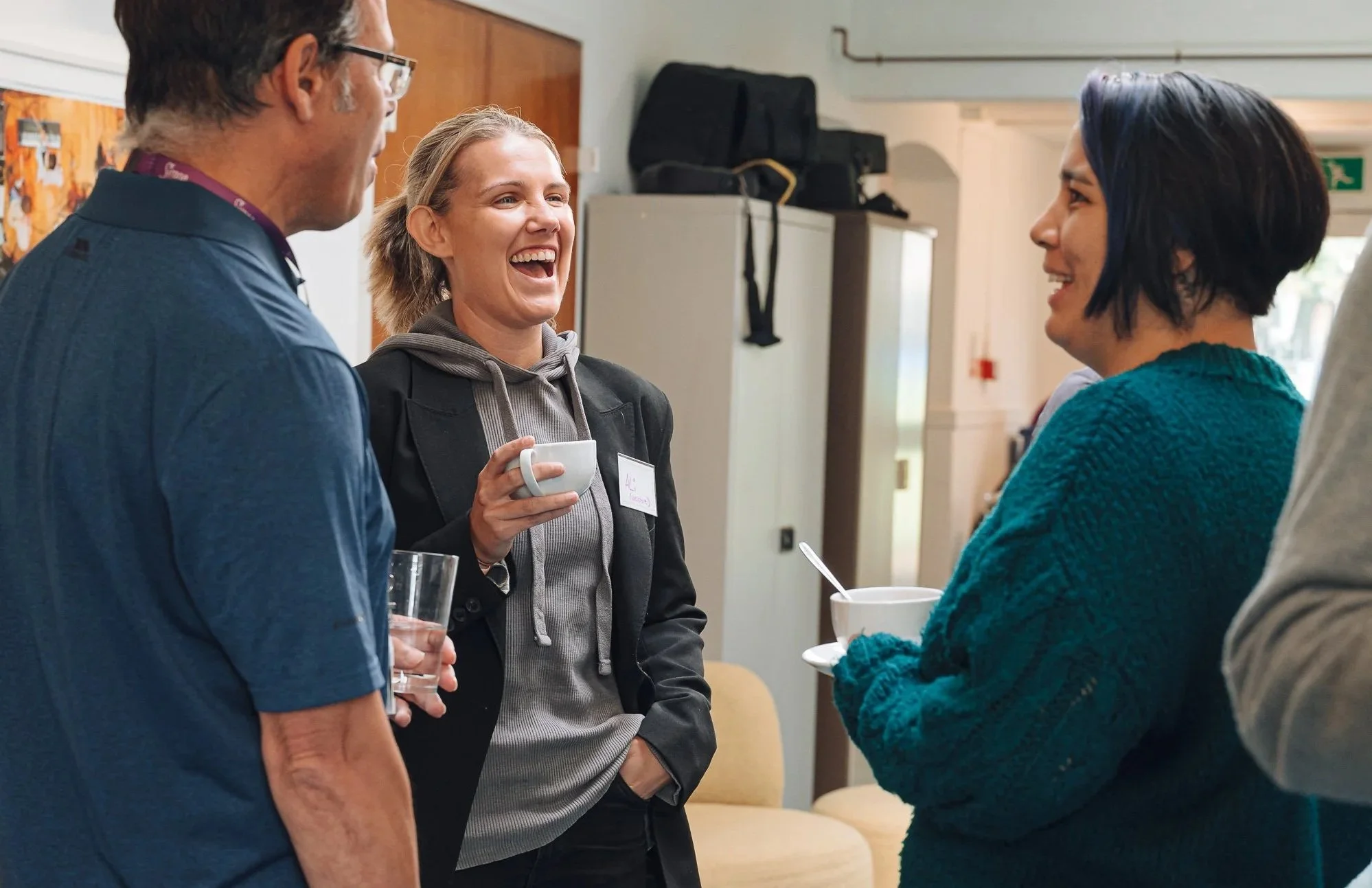 Group of people engaging in conversation, smiling, and holding cups and glasses in an indoor setting.