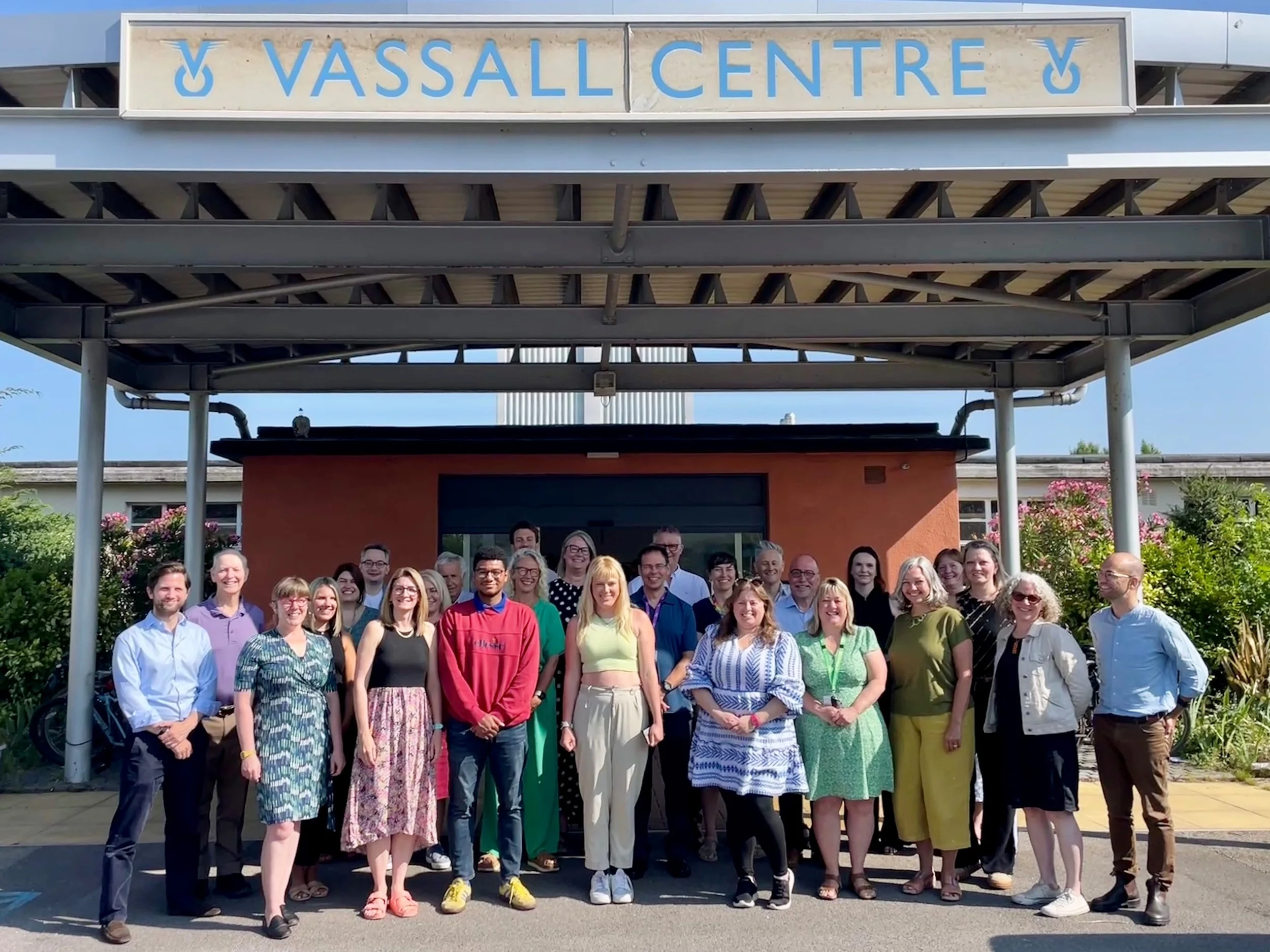 Group of Impact Alliance members standing in front of Vassall Centre entrance, smiling for the photo on a sunny day with greenery around.