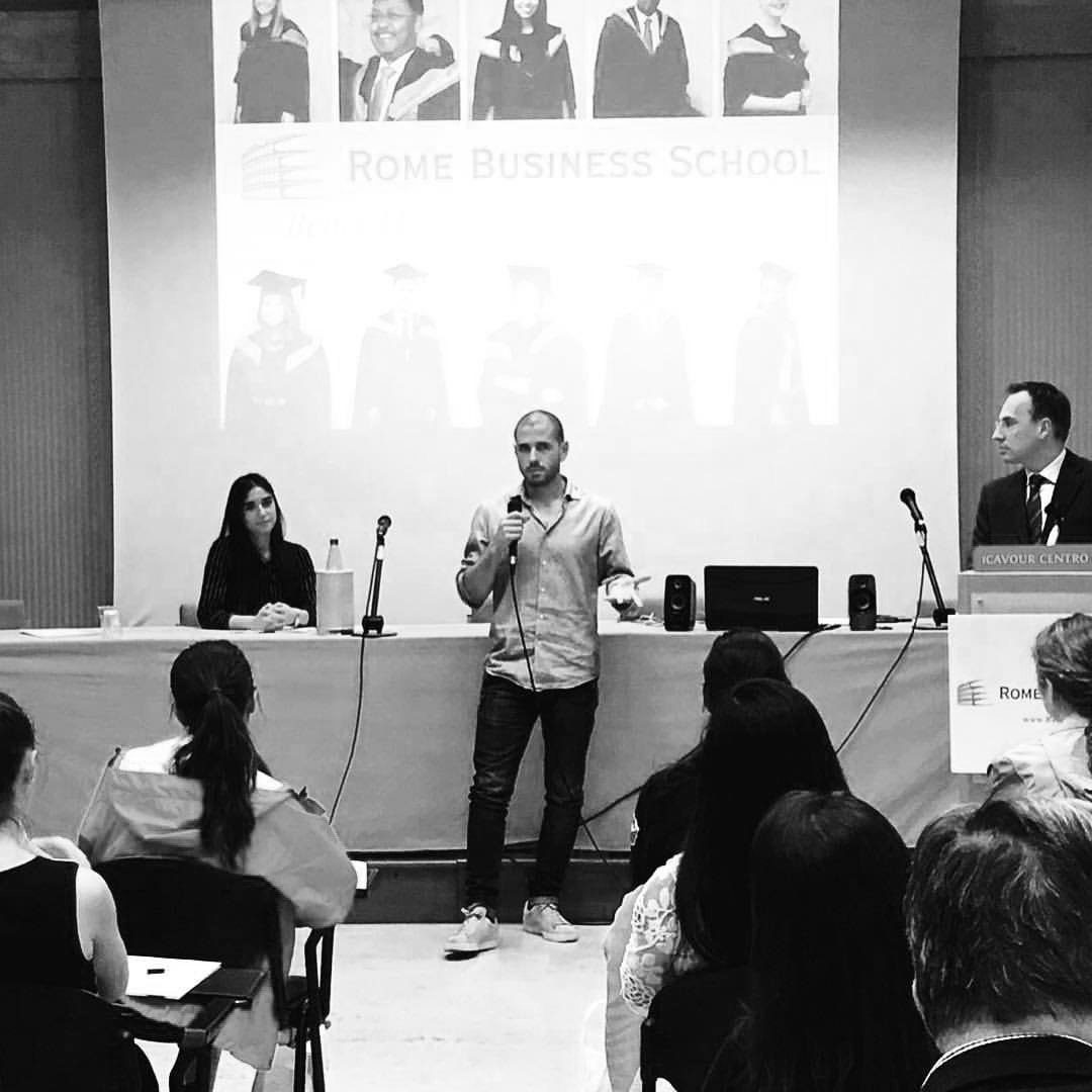 A man is standing and speaking into a microphone in an auditorium, facing an audience of mostly women. Behind him, a large screen displays photographs of people in graduation caps and gowns with the text 'Rome Business School'. To his right, a woman and a man are sitting at a table, the man is at a podium with a sign that reads 'ICAVOUR CENTRO'.