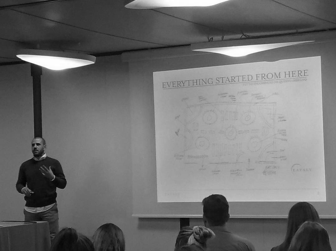 A man giving a presentation in front of a conference room, with an audience. A projected slide titled 'EVERYTHING STARTED FROM HERE' displays a sketch or diagram.