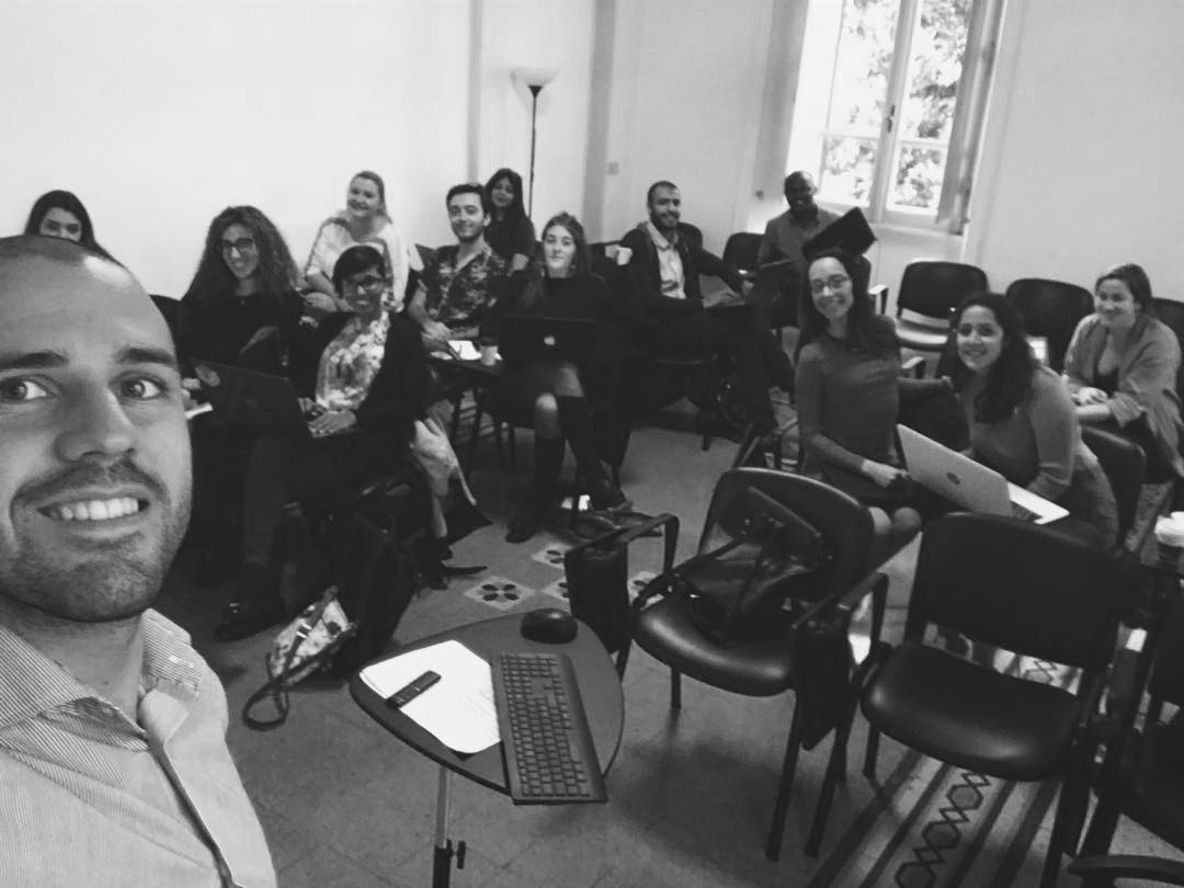 Black and white photo of a man taking a selfie in a conference room with 12 people seated behind him, some with laptops, smiling at the camera, large windows on the right letting in natural light.