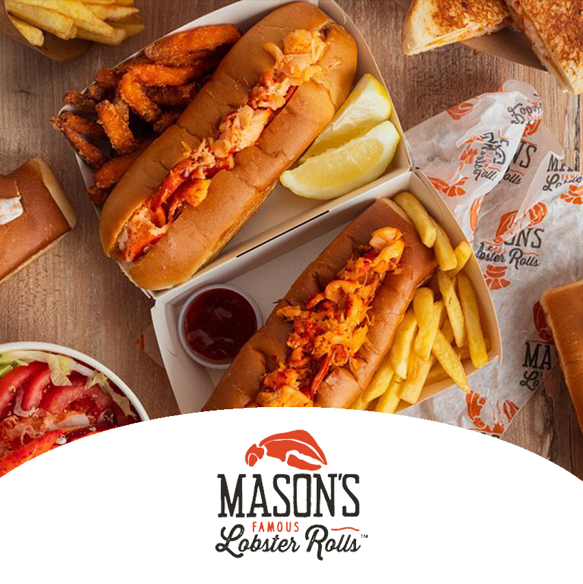 Two lobster rolls with fries and lemon wedges on a table, alongside a small bowl of ketchup, and other food items in the background, with a Mason's Famous Lobster Rolls logo at the bottom.