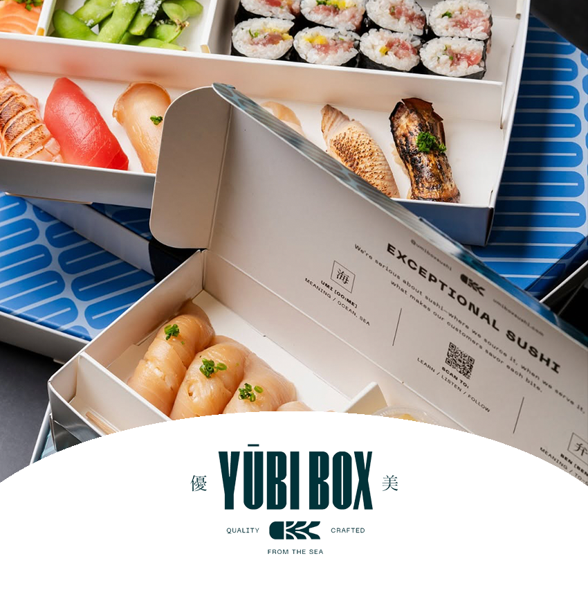 Open sushi boxes containing various sushi pieces, including nigiri with fish, sushi rolls, and sashimi, on a blue patterned surface.