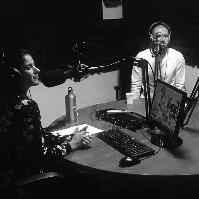 Two people in a radio recording studio with microphones, headphones, a computer monitor, and miscellaneous equipment on a round table.