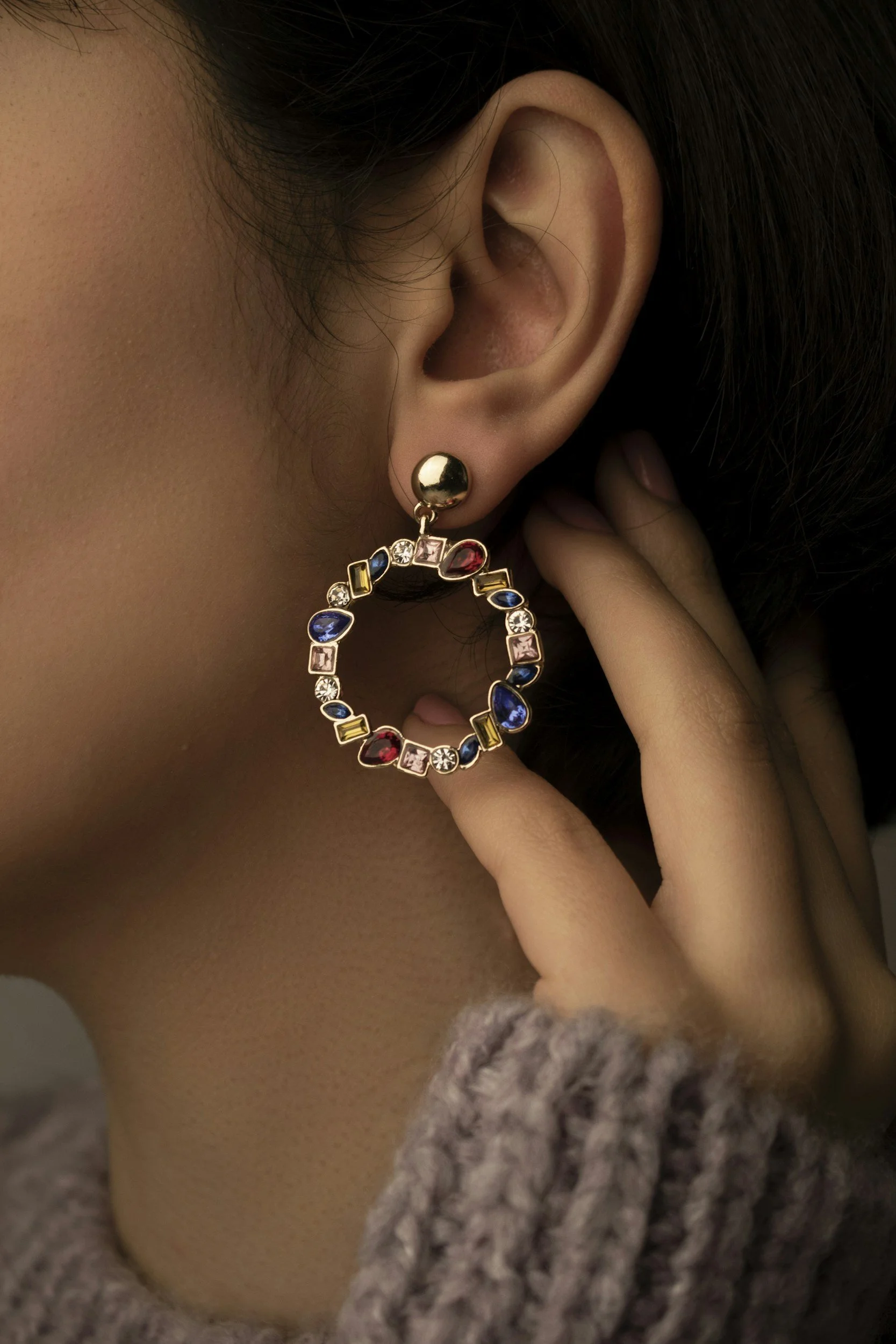 Close-up of a person's ear wearing a colorful, jeweled hoop earring, with part of their face and hand visible, and wearing a soft, knit sweater.