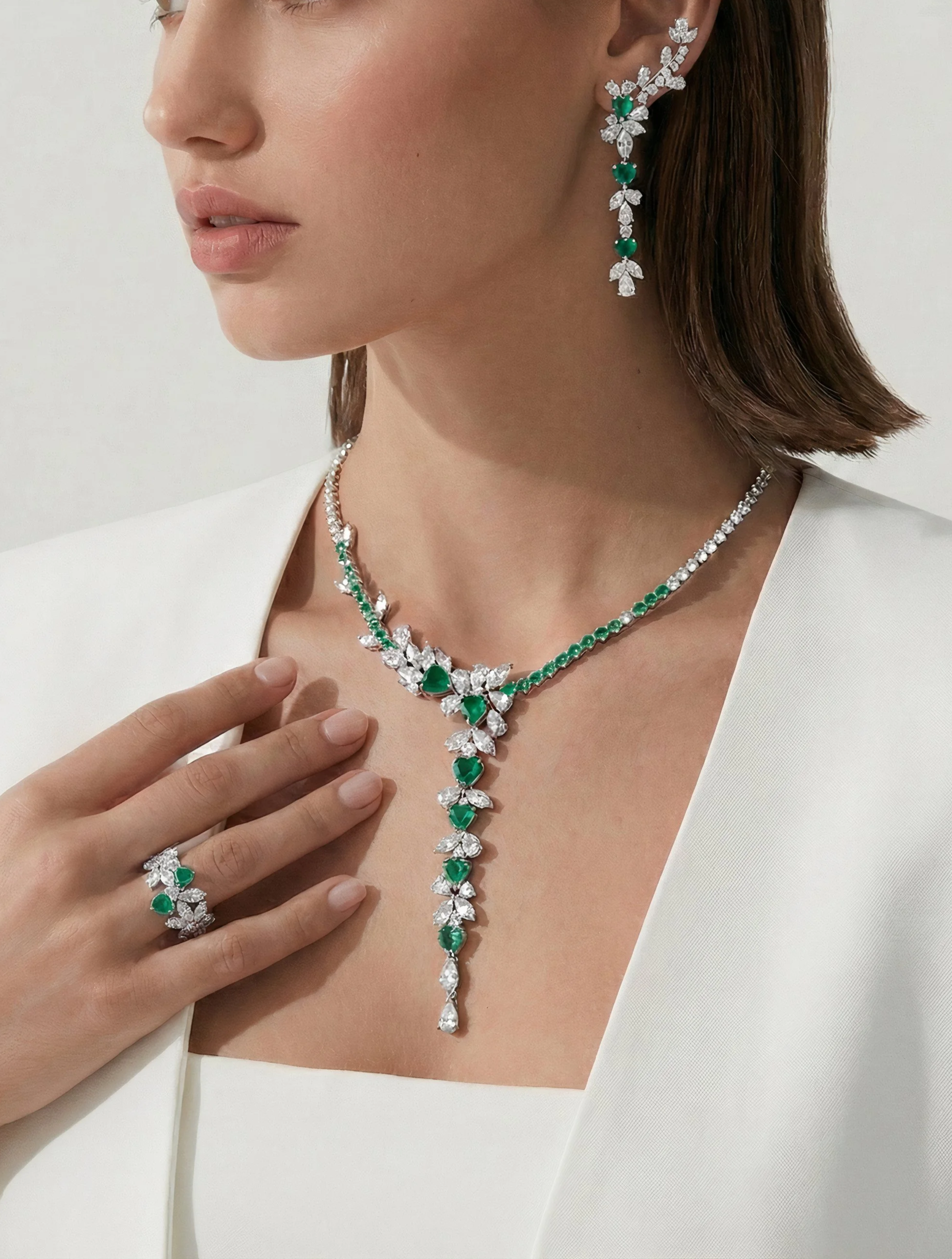 Close-up of a woman wearing a white blazer, adorned with a matching jewelry set that includes earrings, a necklace, a ring, and a bracelet, all featuring green gemstones and diamonds.