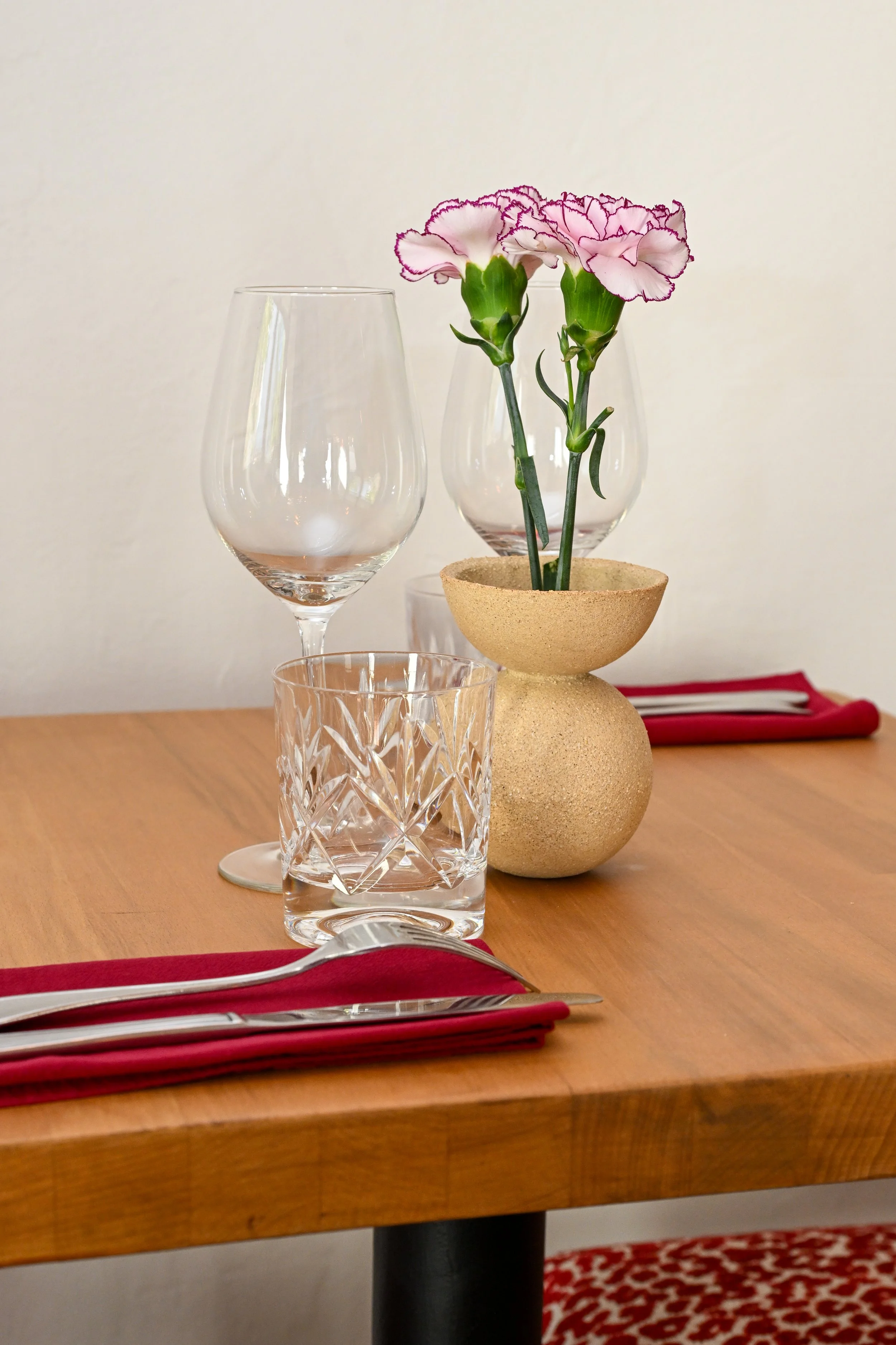 Une table en bois avec un verre à vin vide, un verre en cristal, un couteau et une fourchette avec un torchon rouge en dessous, une plante avec deux fleurs roses dans un vase beige en forme de tête d'animal, et en arrière-plan, un autre vase transparent vide, un tissu rouge et blanc avec un motif animal