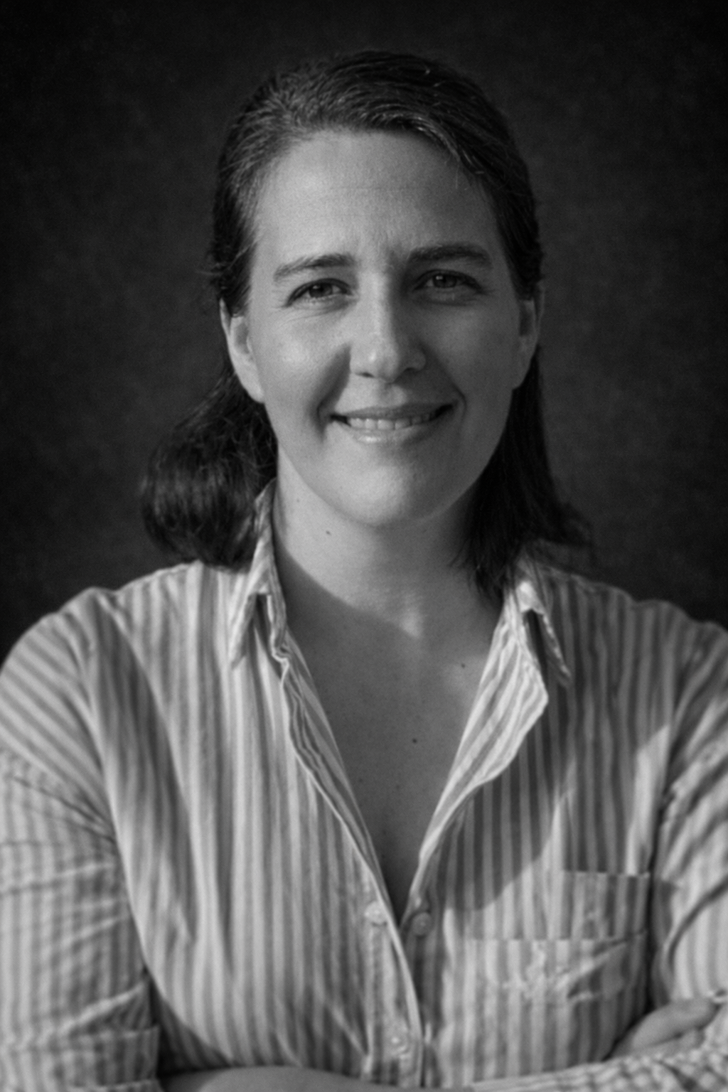 Kadın, pembe ve beyaz çizgili gömlek giymiş, kolunu çapraz yapmış, gülümseyerek duruyor.