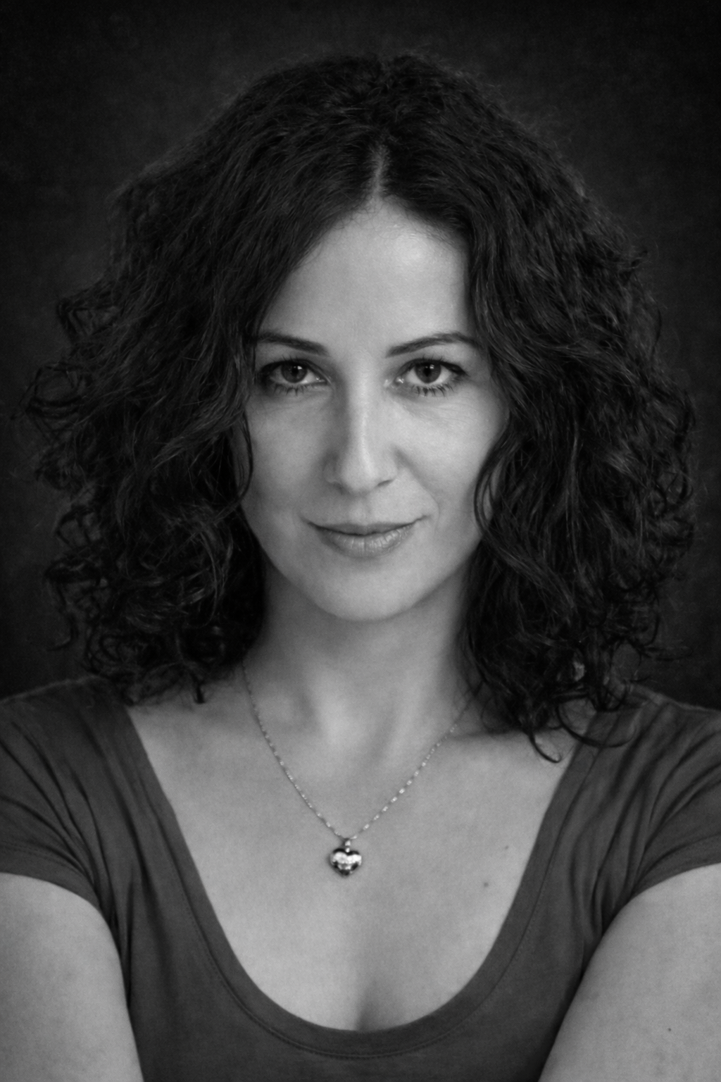 Kahverengi, dalgalı saçlara sahip kadın kısa kollu kırmızı tişört giymiş ve gümüş bir kolye takmış, arka plan siyah ve gri geçişlidir.