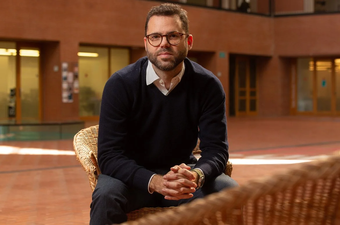 Un home amb barba, ulleres i roba formal, assegut en una cadira de vímet en un espai interior amb parets de maó.